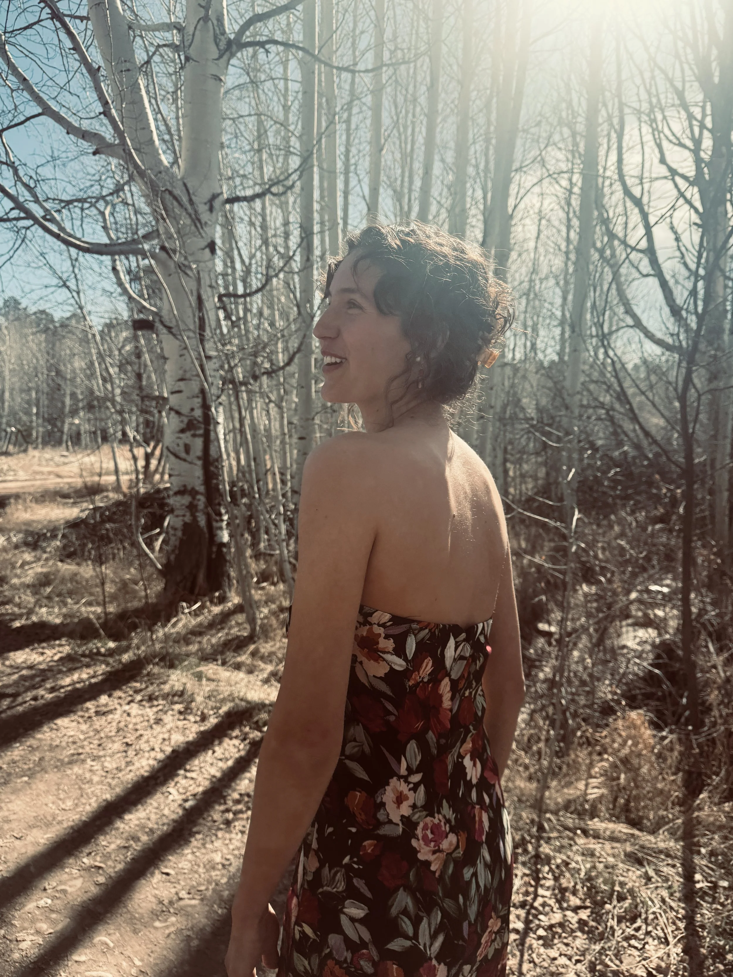 A woman with short curly hair, wearing a strapless floral dress, standing in a forest with leafless trees, sunlight shining through, and shadows on the ground.