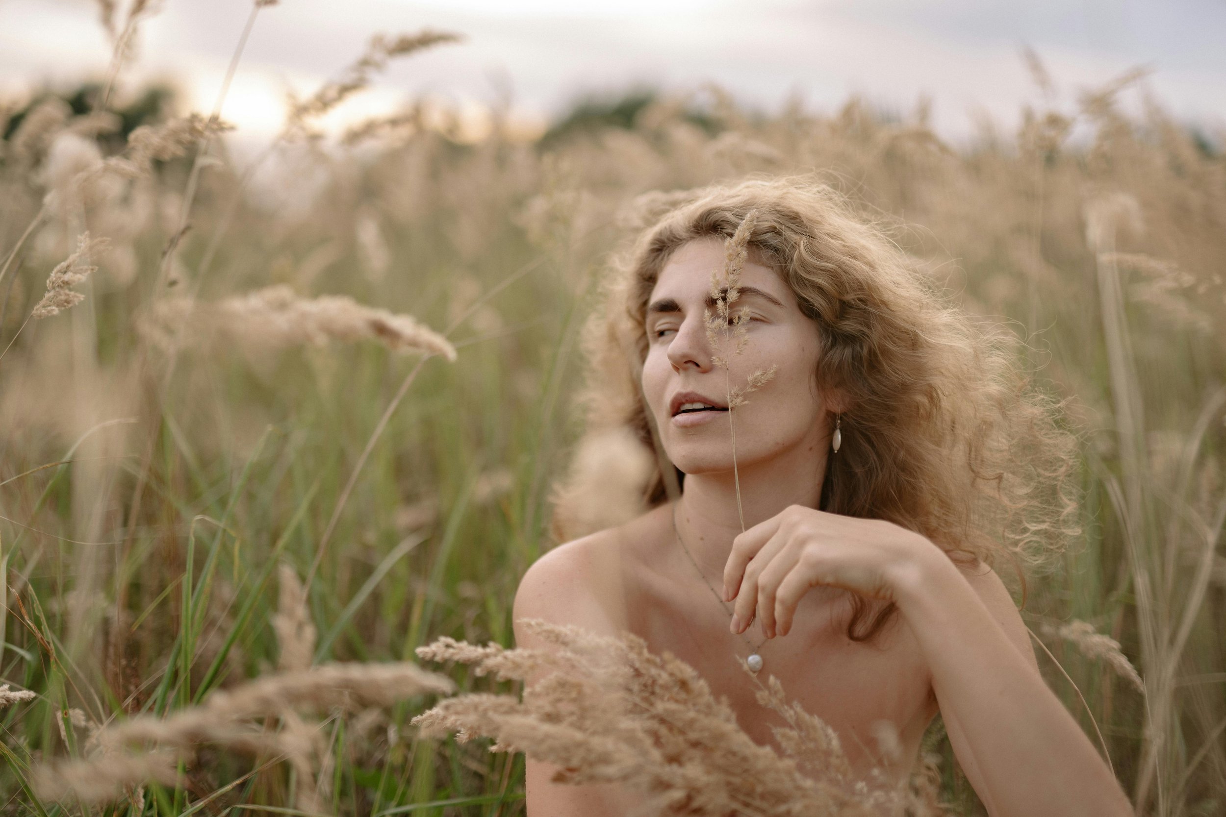 A woman with curly hair sitting in a field of tall grass during dusk.