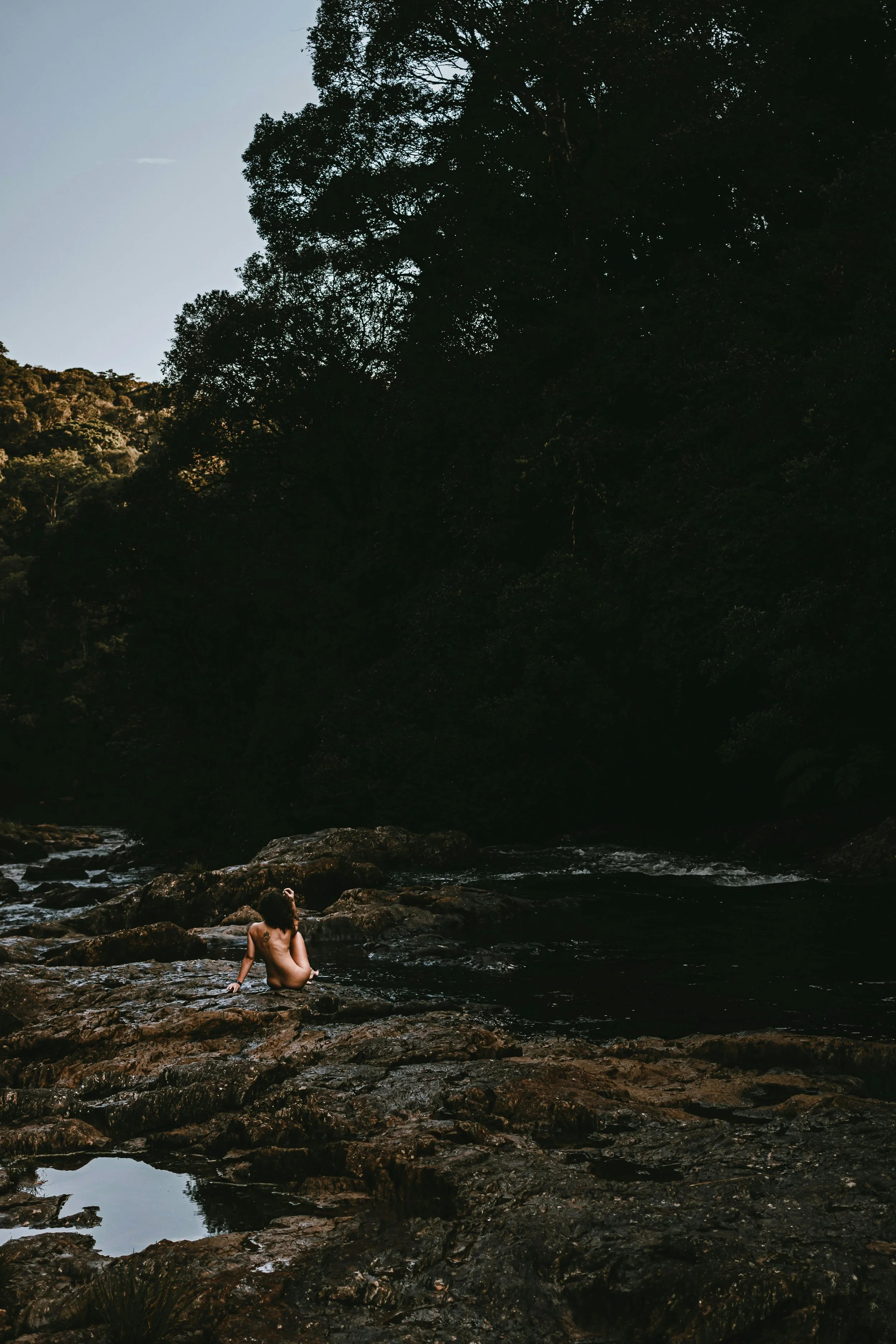 A woman sitting on a rocky riverbank, with a background of dense trees and a darkening sky.