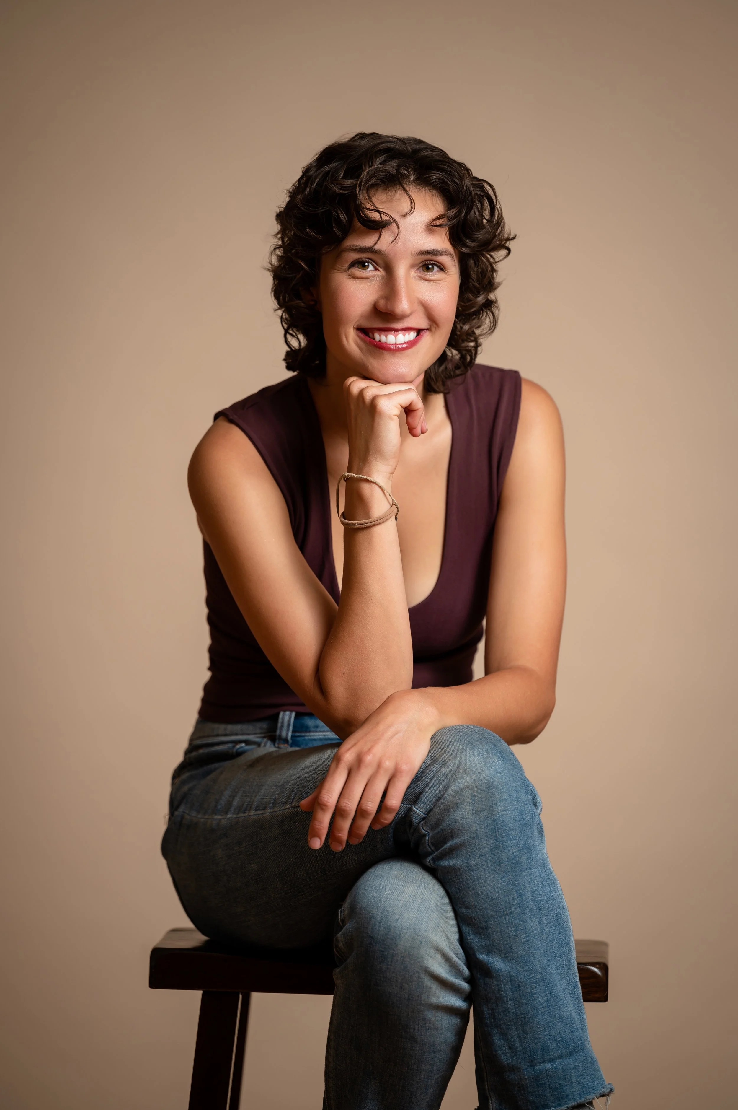 A woman with short curly brown hair, wearing a sleeveless maroon top and jeans, sitting on a chair with her hand resting on her chin and smiling at the camera against a plain beige background.