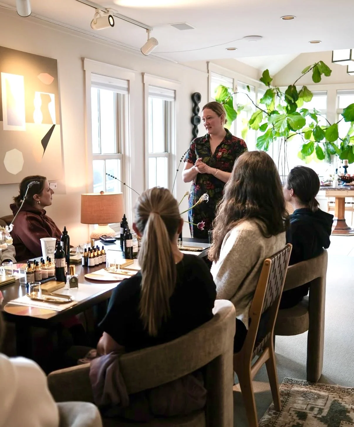 A woman stands and teaches a group of women seated around a dining table indoors. The table has various bottles and makeup items on it. The room is well-lit with natural light from large windows, and there are decorative art pieces and a large green