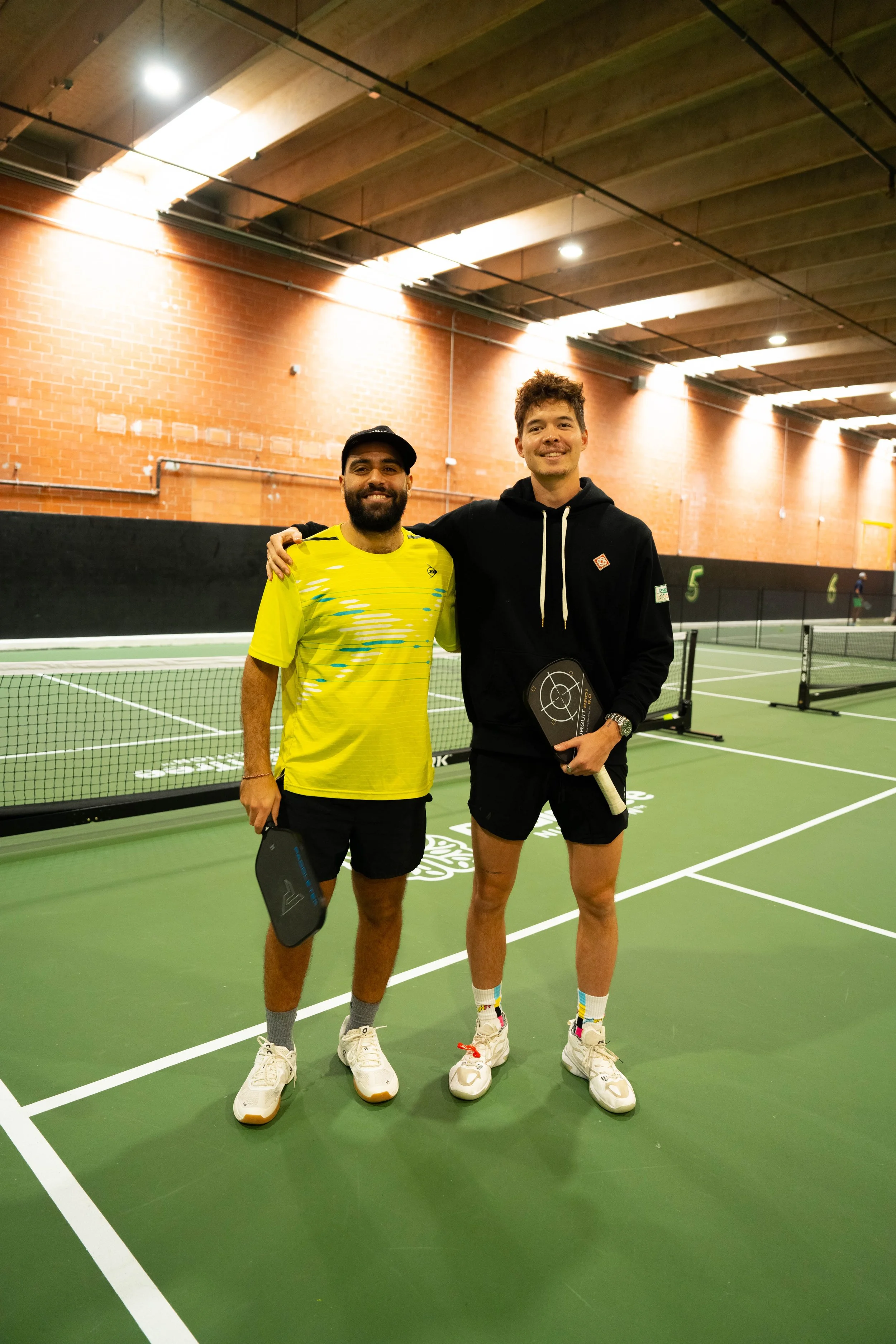 Two men standing on an indoor pickleball court, holding paddles. One is wearing a yellow shirt, and the other a black hoodie.