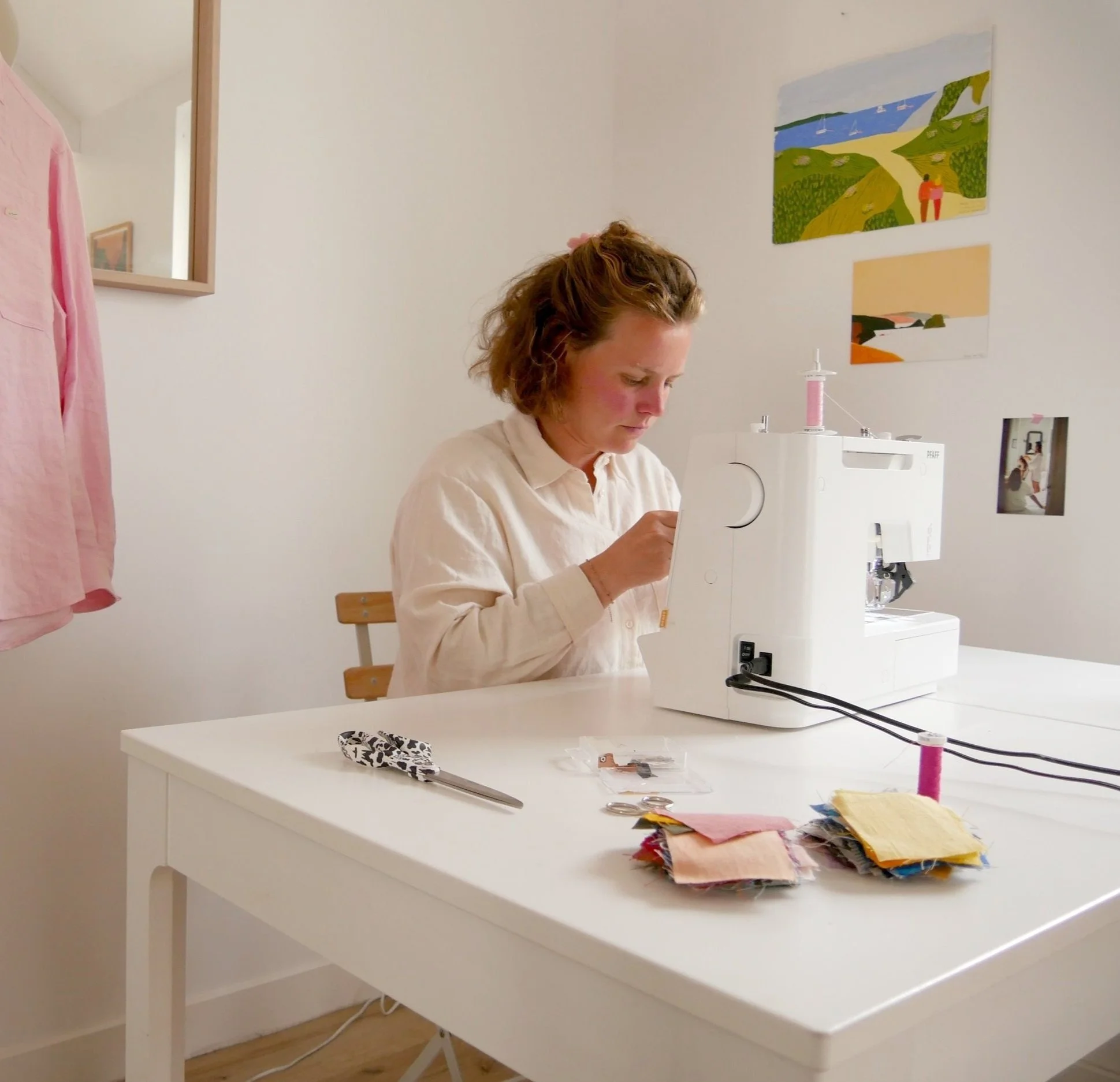 Femme utilisant une machine à coudre avec des échantillons de tissu sur une table blanche dans une pièce lumineuse avec des peintures sur le mur.