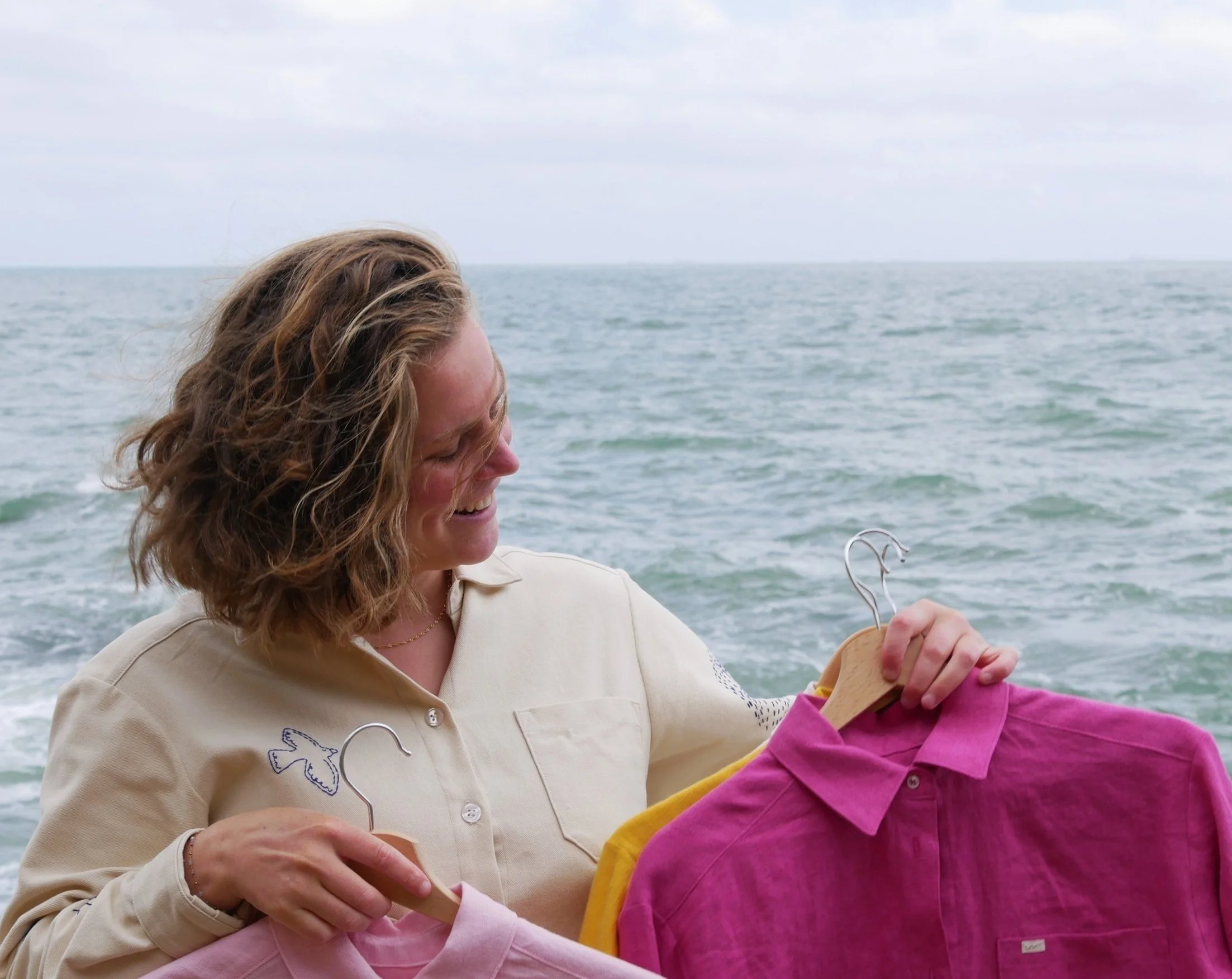 Une femme souriante tenant des vêtements colorés sur des cintres devant la mer.