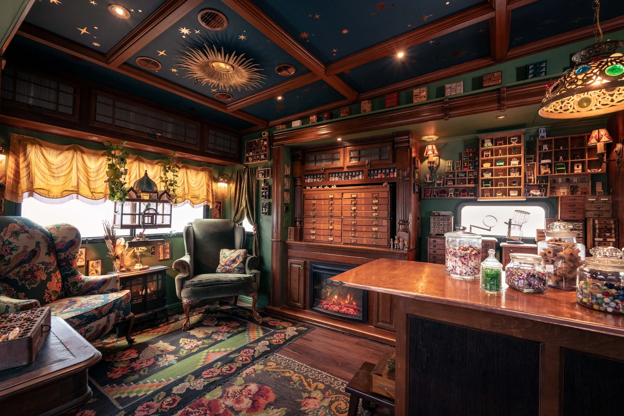 A cozy vintage living room with dark wooden shelves filled with miniature furniture, toys, and collectibles, green walls, floral upholstered furniture, a colorful patterned rug, and a ceiling decorated with gold stars and a sunburst light fixture.