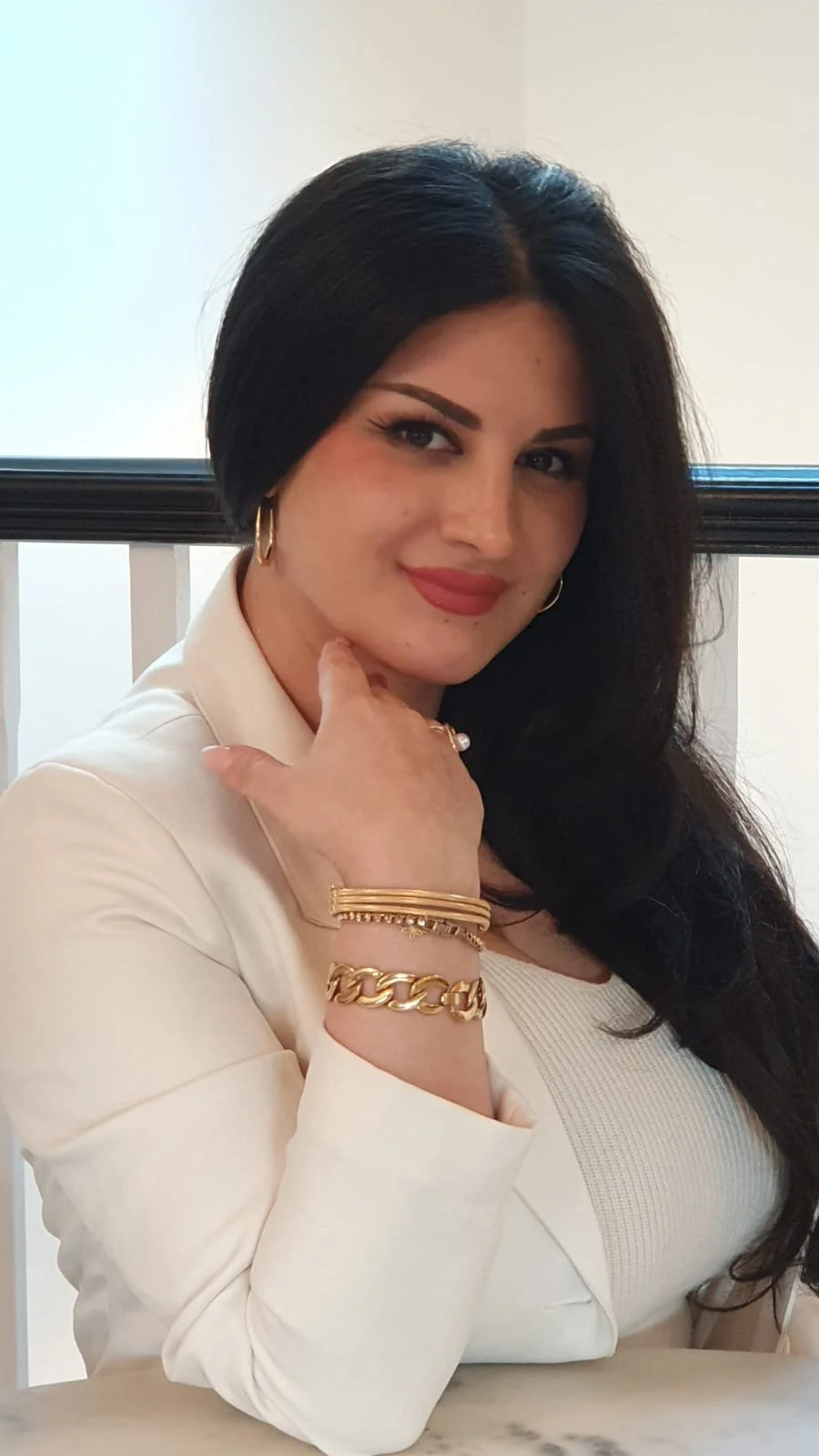A woman with long dark hair and light makeup sitting at a table, wearing a cream-colored blazer and gold jewelry, smiling at the camera.