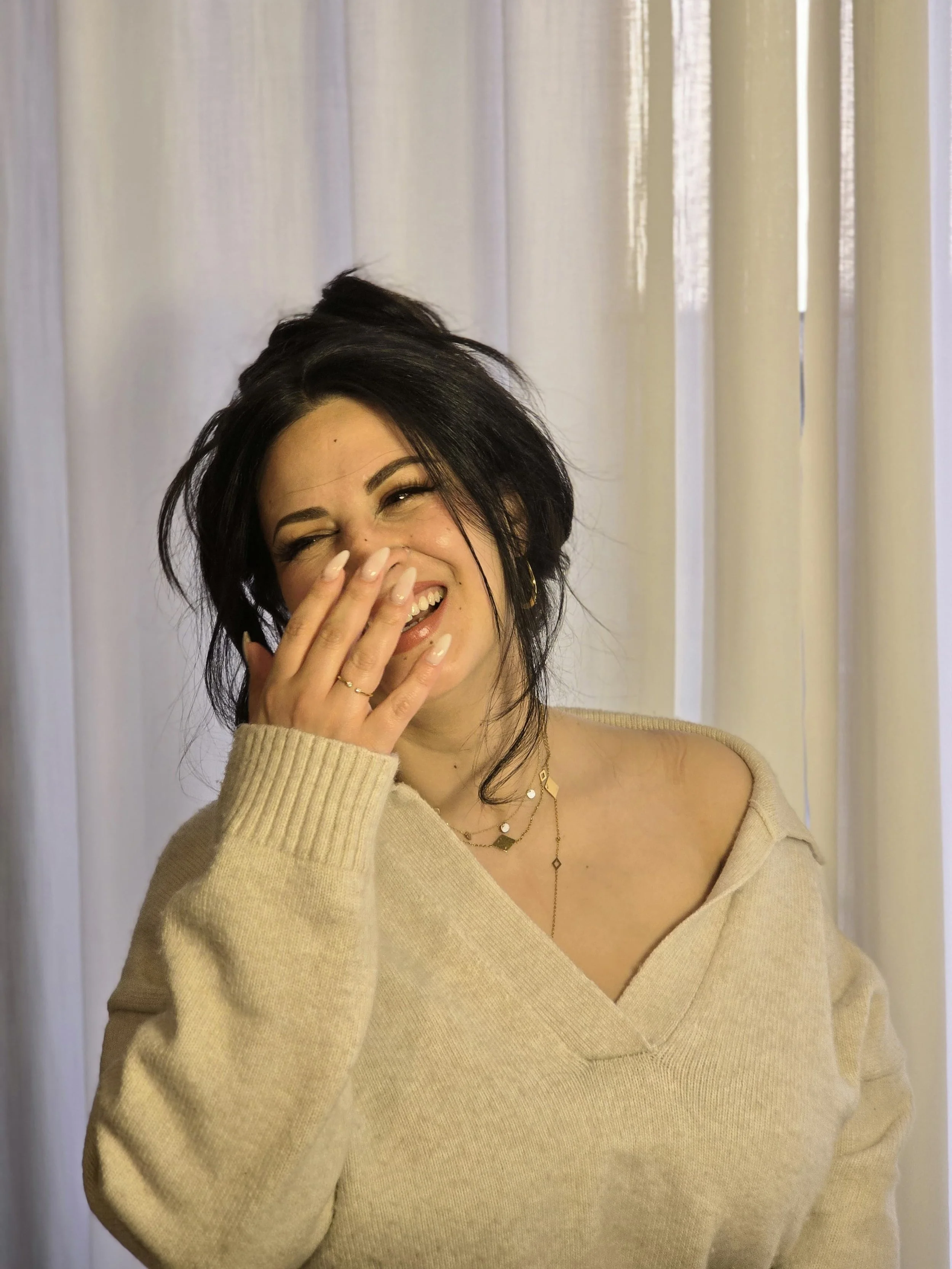 Life Coach Lama - A woman with long black hair, loosely and effortlessly tied up. Soft, fuzzy, white jumper, soft lighting, lots of gold jewellery. Lama is se laughing and covering her mouth with her hand, standing in front of light-colored curtains.