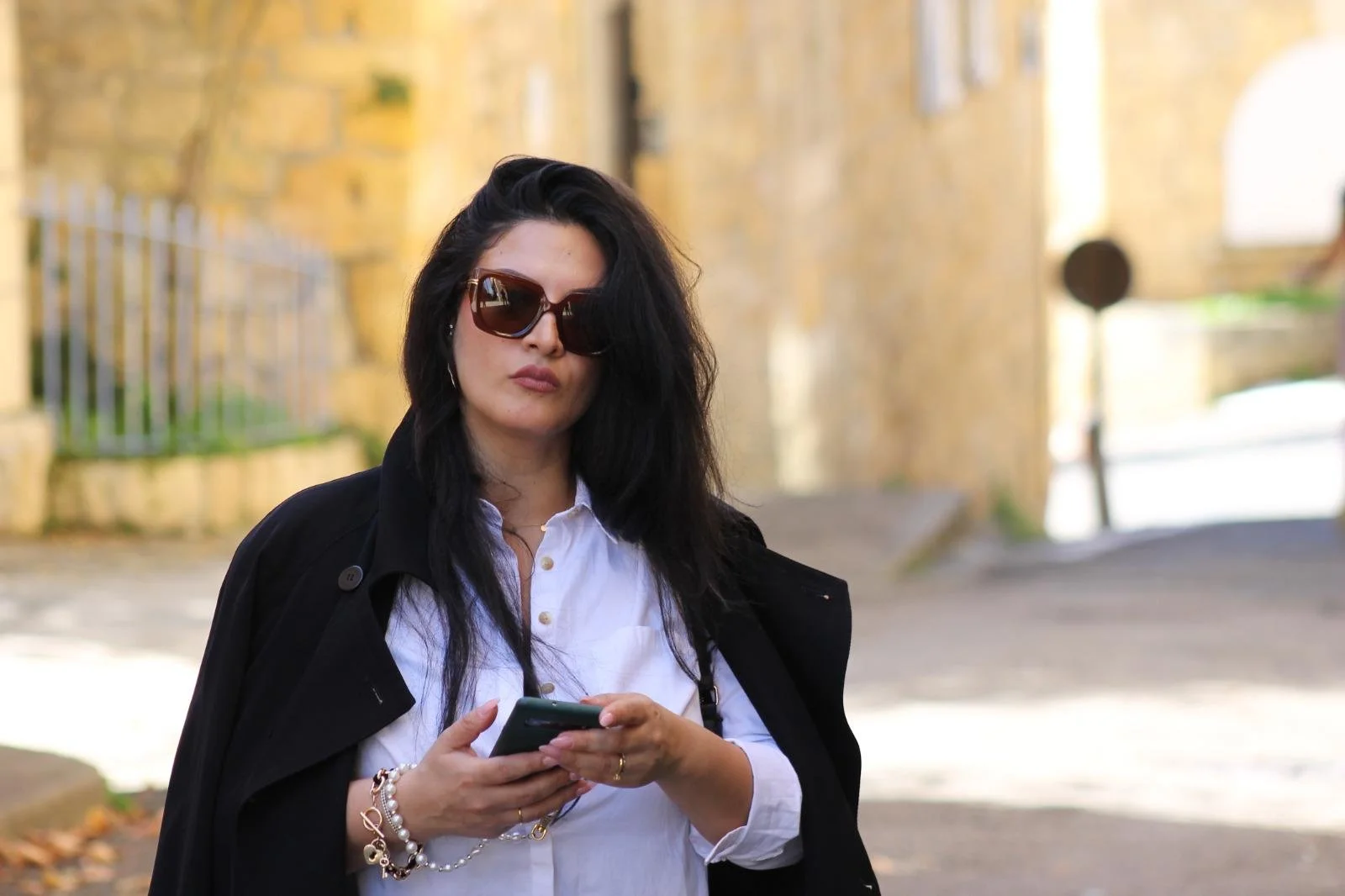 Life Coach Lama seen in candid shot, dark hair and sunglasses holding a phone while walking outdoors on a city street with yellow brick buildings in the background.