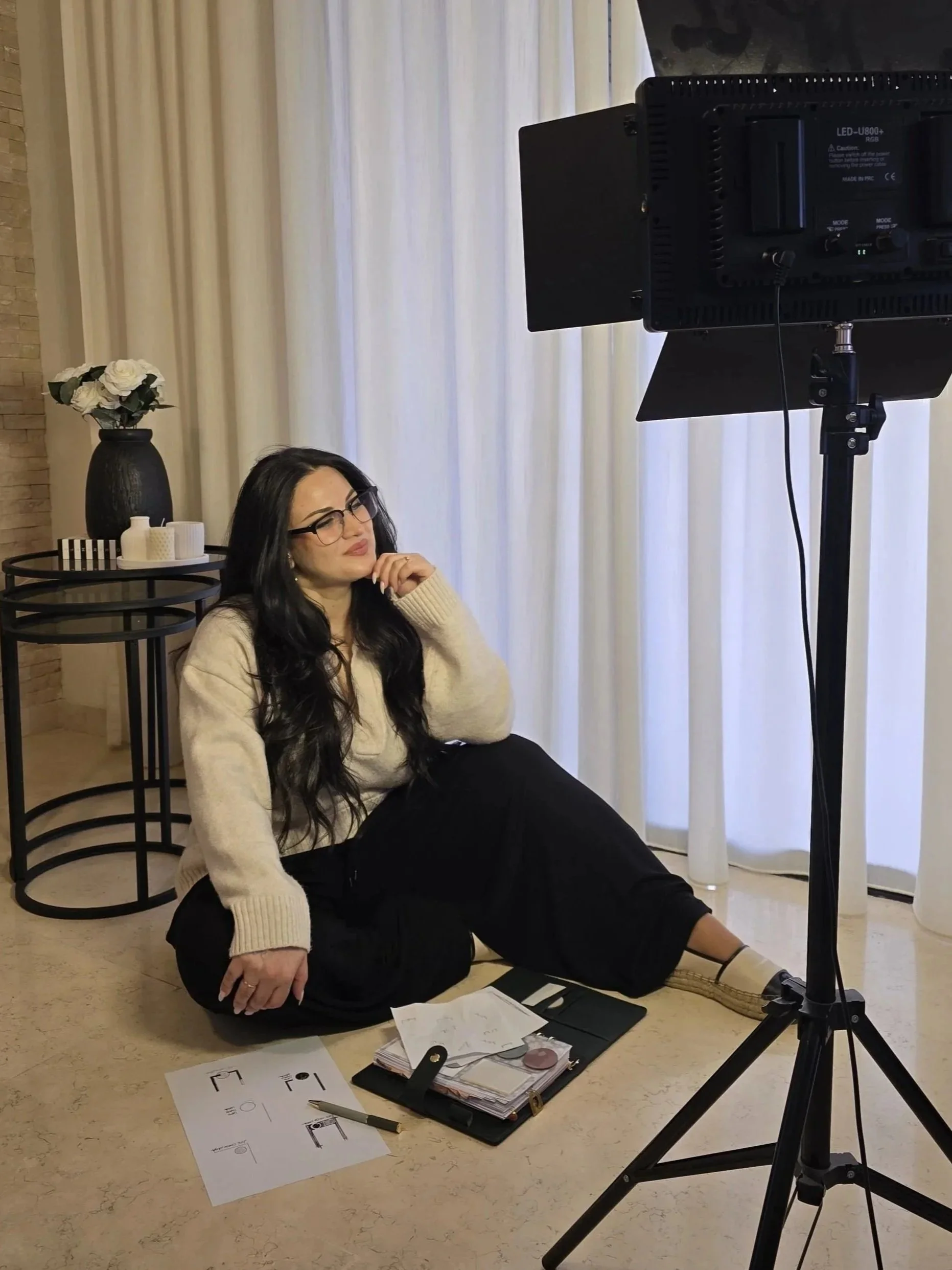Life Coach Lama-woman with long black hair, glasses, wearing a beige jumper and black pants, sitting on the floor in front of a camera, with old logos & notebooks scattered around, in a room with white curtains and a side table with decorative items.