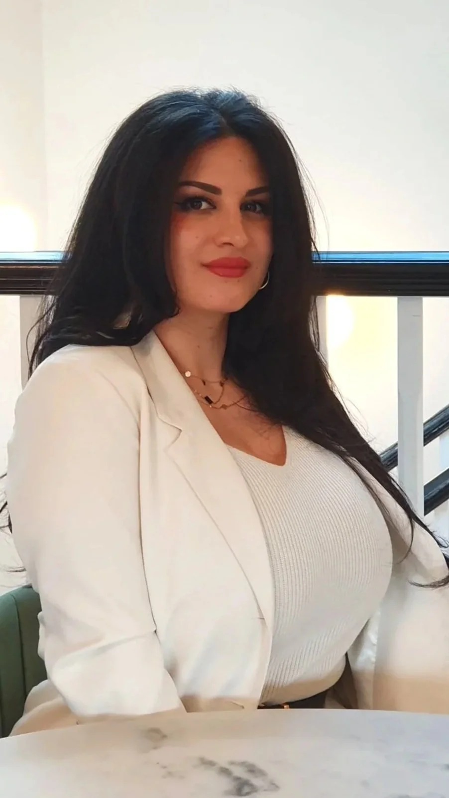 A woman with long dark hair, wearing a white blazer and beige top, sitting indoors near a window with black trim.