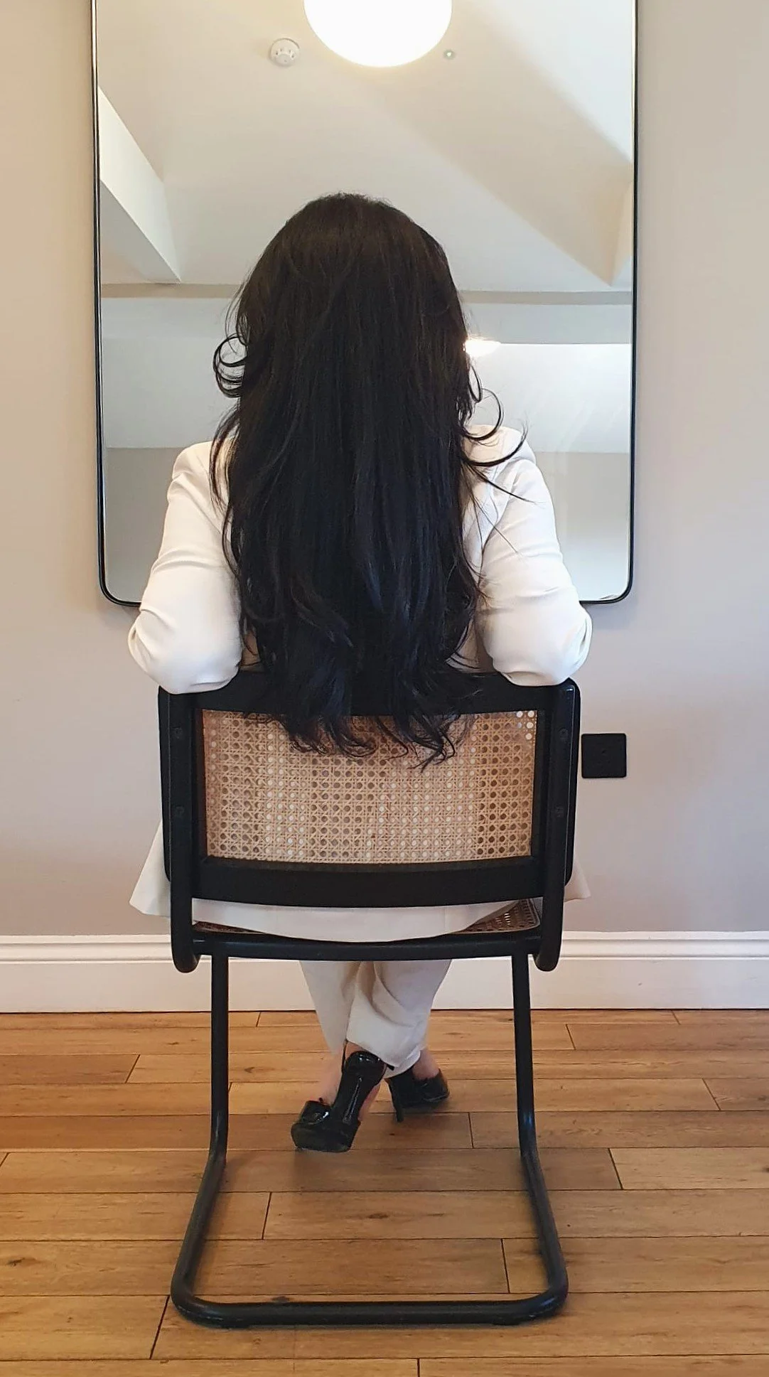 A woman with long black hair sitting in a chair, facing a mirror on a light-colored wall. She is wearing a white blazer and beige pants, in a room with wooden flooring and modern decor.