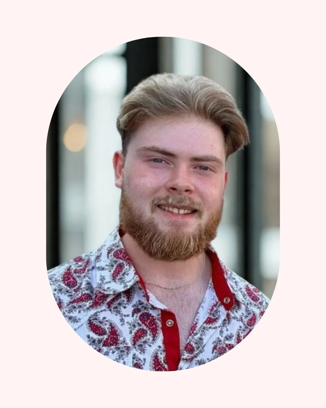 Portrait of a young man with light brown hair and a beard, smiling, wearing a patterned button-up shirt, outdoors during daytime.