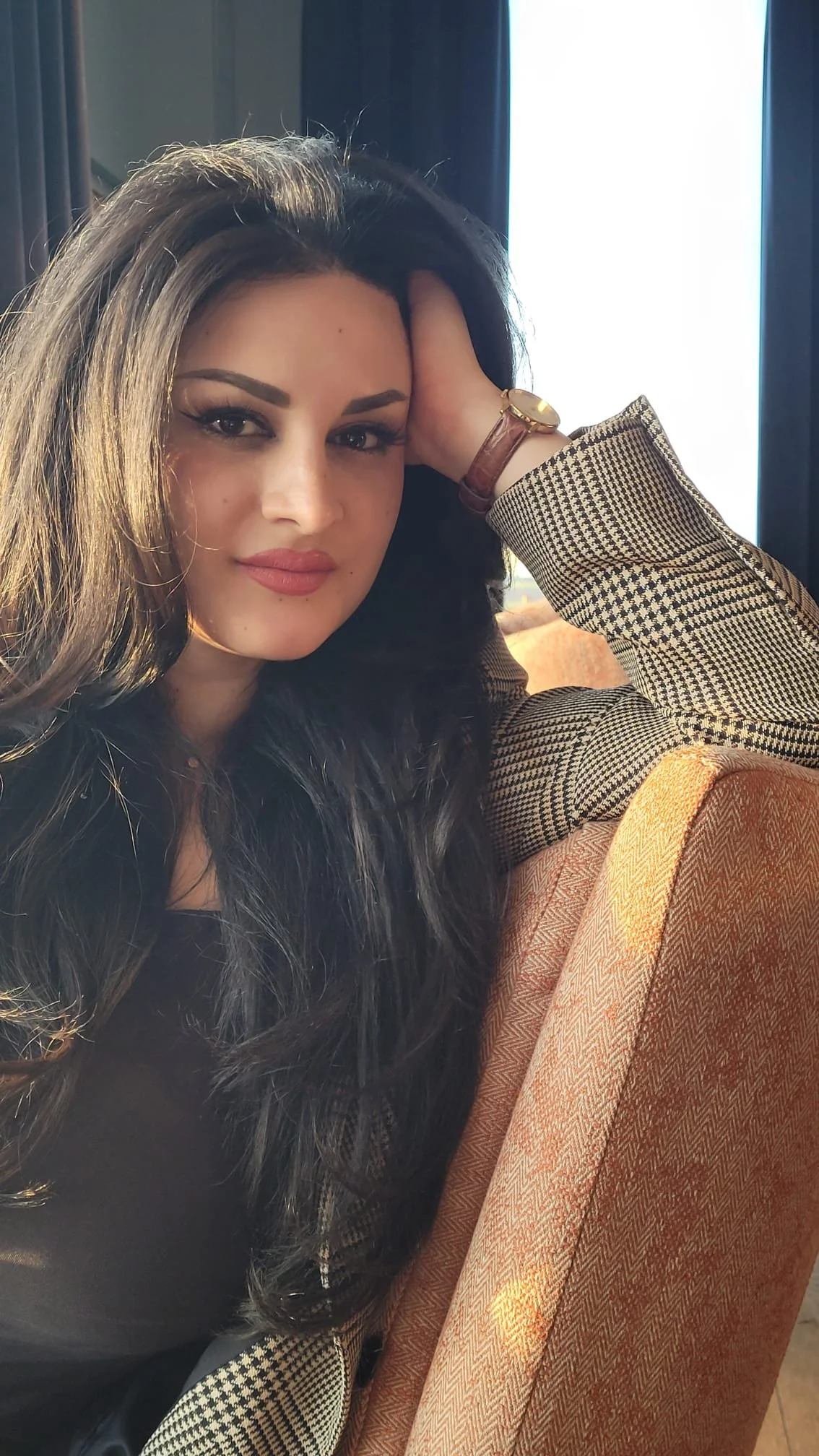 A woman with long dark hair sitting on an orange upholstered chair near a window, wearing a patterned jacket, with her hand resting on her head, looking at the camera.