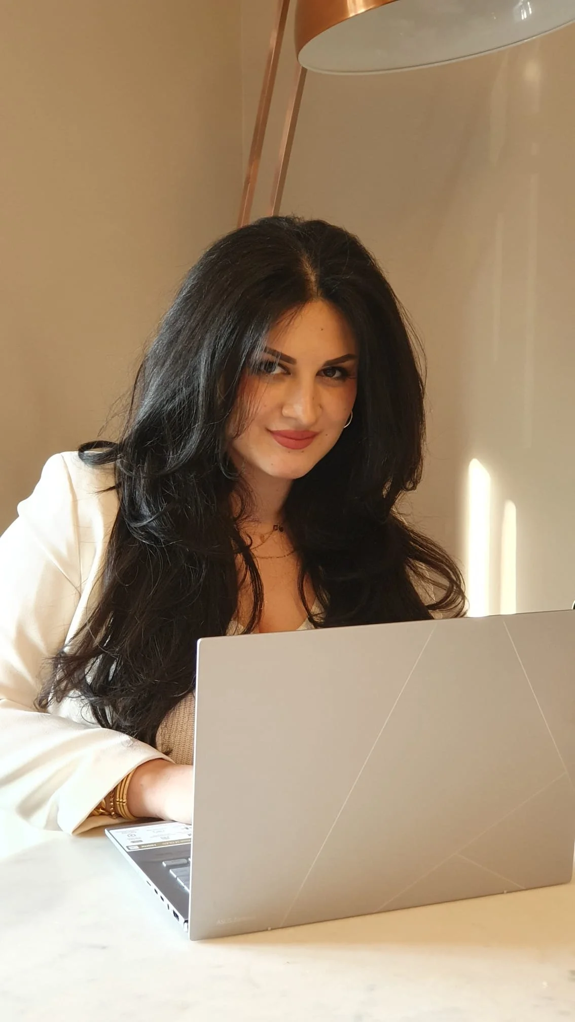 A woman with long black hair sitting at a table with a silver laptop in front of her. She is smiling slightly and looking at the camera. The background features a beige wall and a copper-colored lamp.