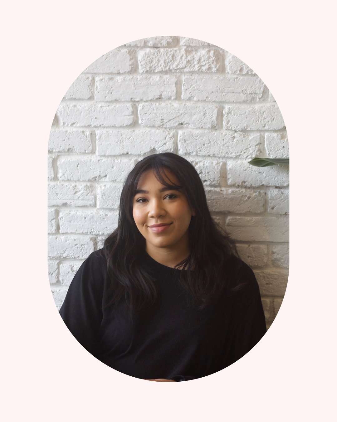 A young woman with dark hair and a black shirt smiling against a white brick wall. Life Coach Lama client success - Life Coach London - Lama Abou Daher