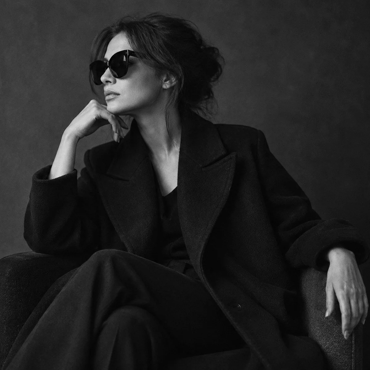Woman in a black coat and sunglasses seated in an armchair, in black and white