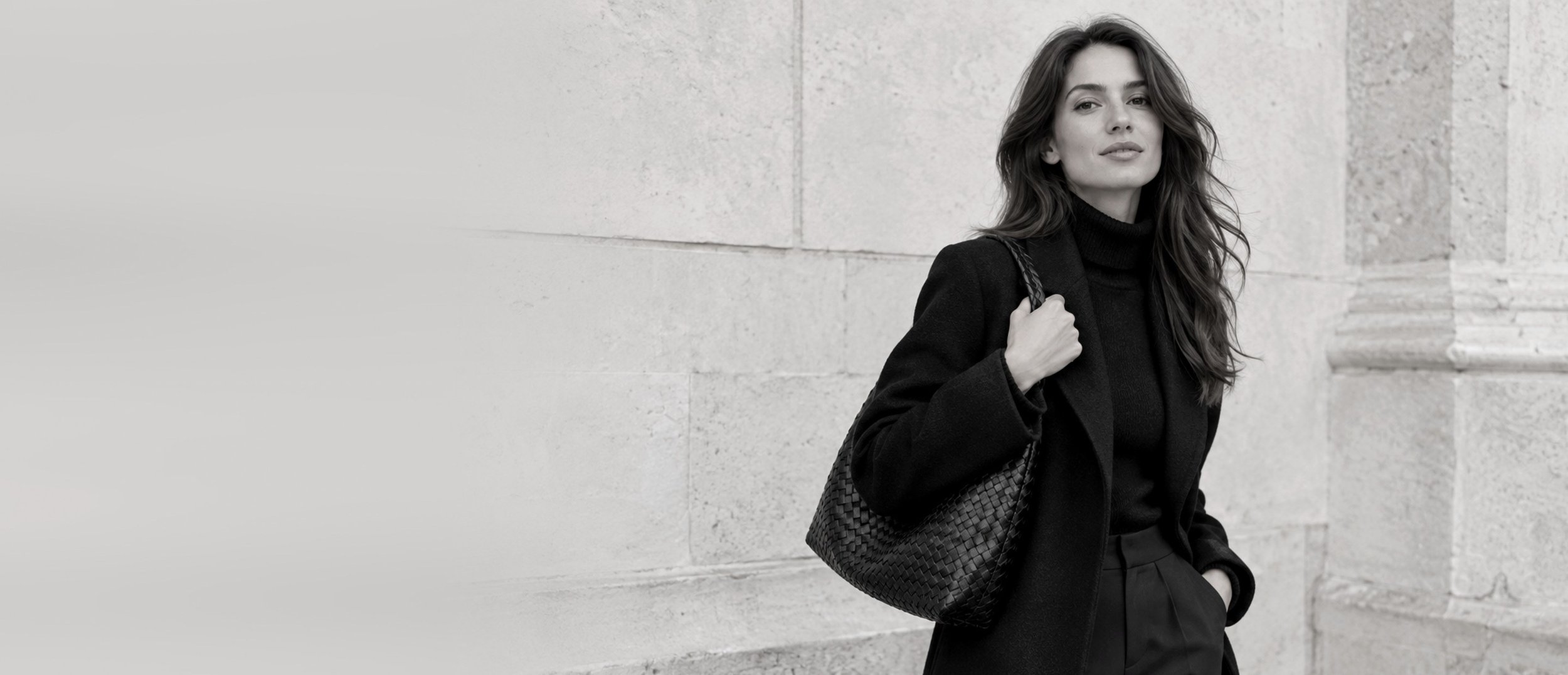 Woman in a long black coat with a woven black shoulder bag beside a pale stone wall, in black and white