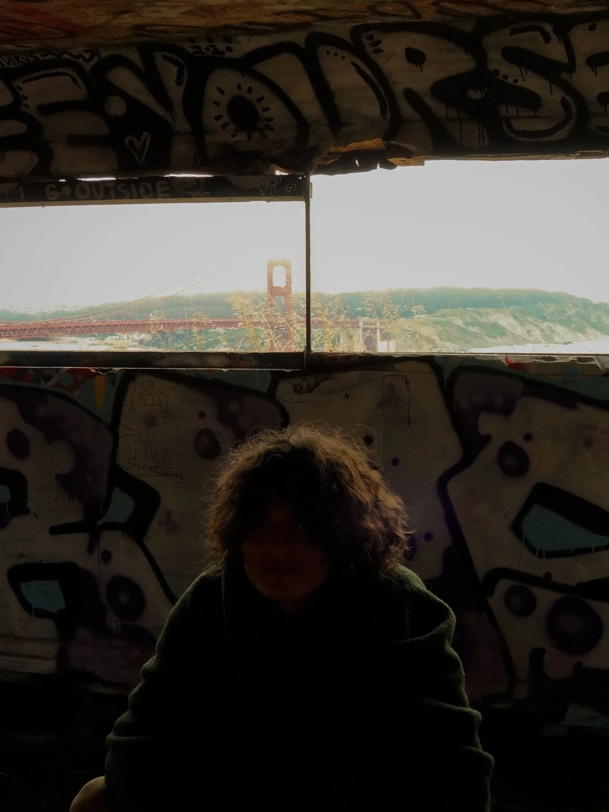 A person with curly hair sitting inside a graffiti-covered structure with a window, overlooking the Golden Gate Bridge in San Francisco, California.