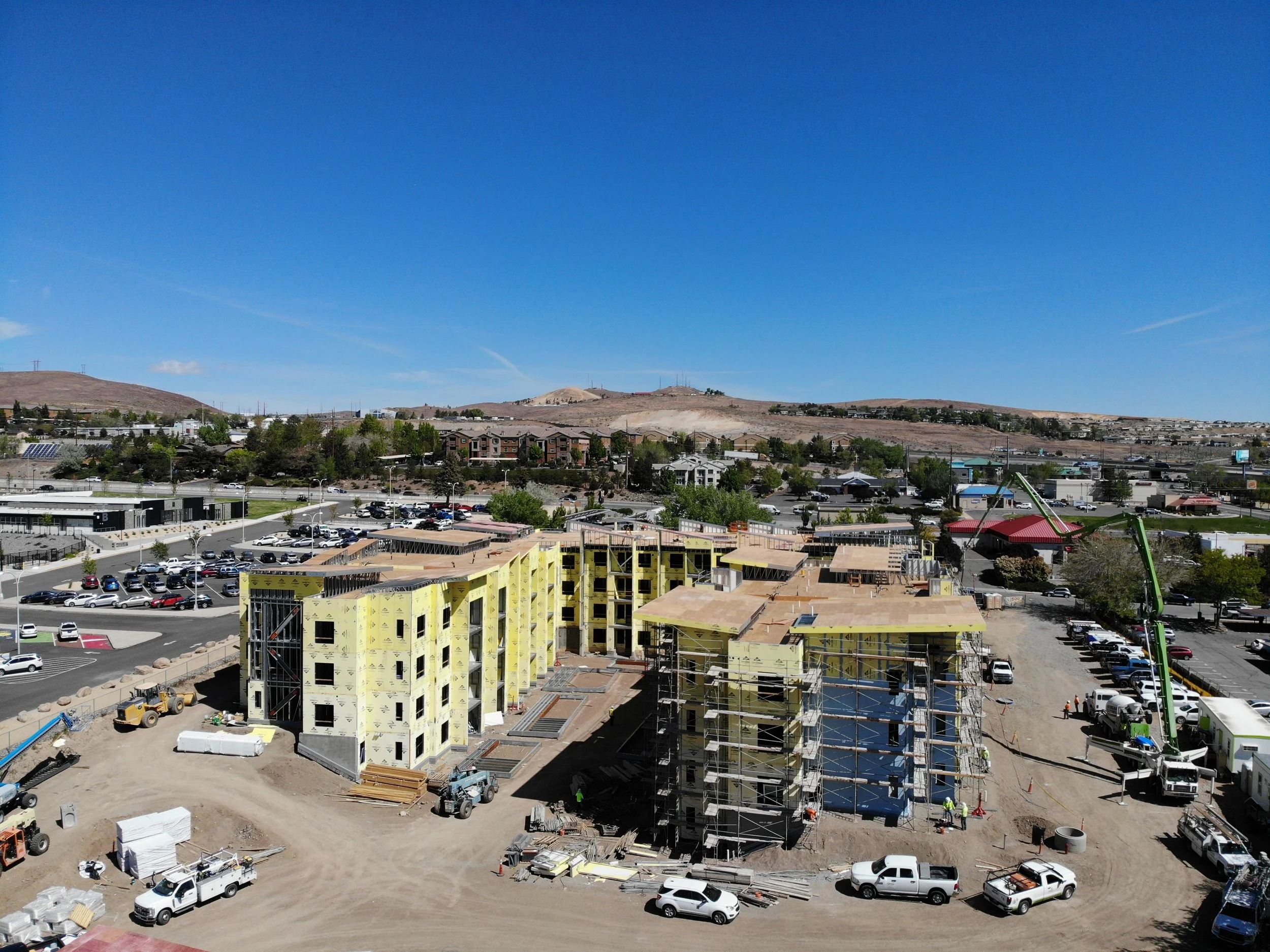 Construction site with trucks, conveyors, and a yellow excavator spreading dirt, surrounded by parked vehicles and a nearby shopping area with buildings and parked cars.