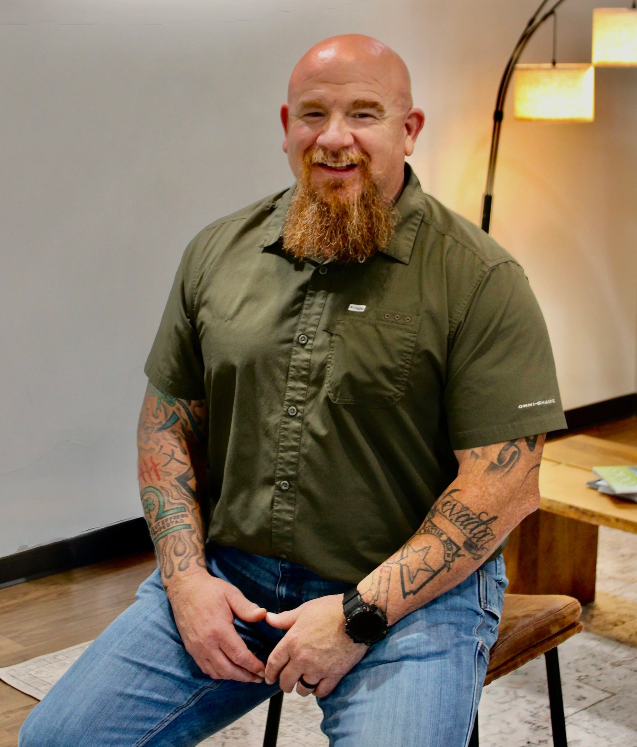 A smiling, bald man with a red beard and tattoos on his arms, wearing a green short-sleeved shirt and blue jeans, sitting on a stool in a room with a floor lamp and wooden furniture.