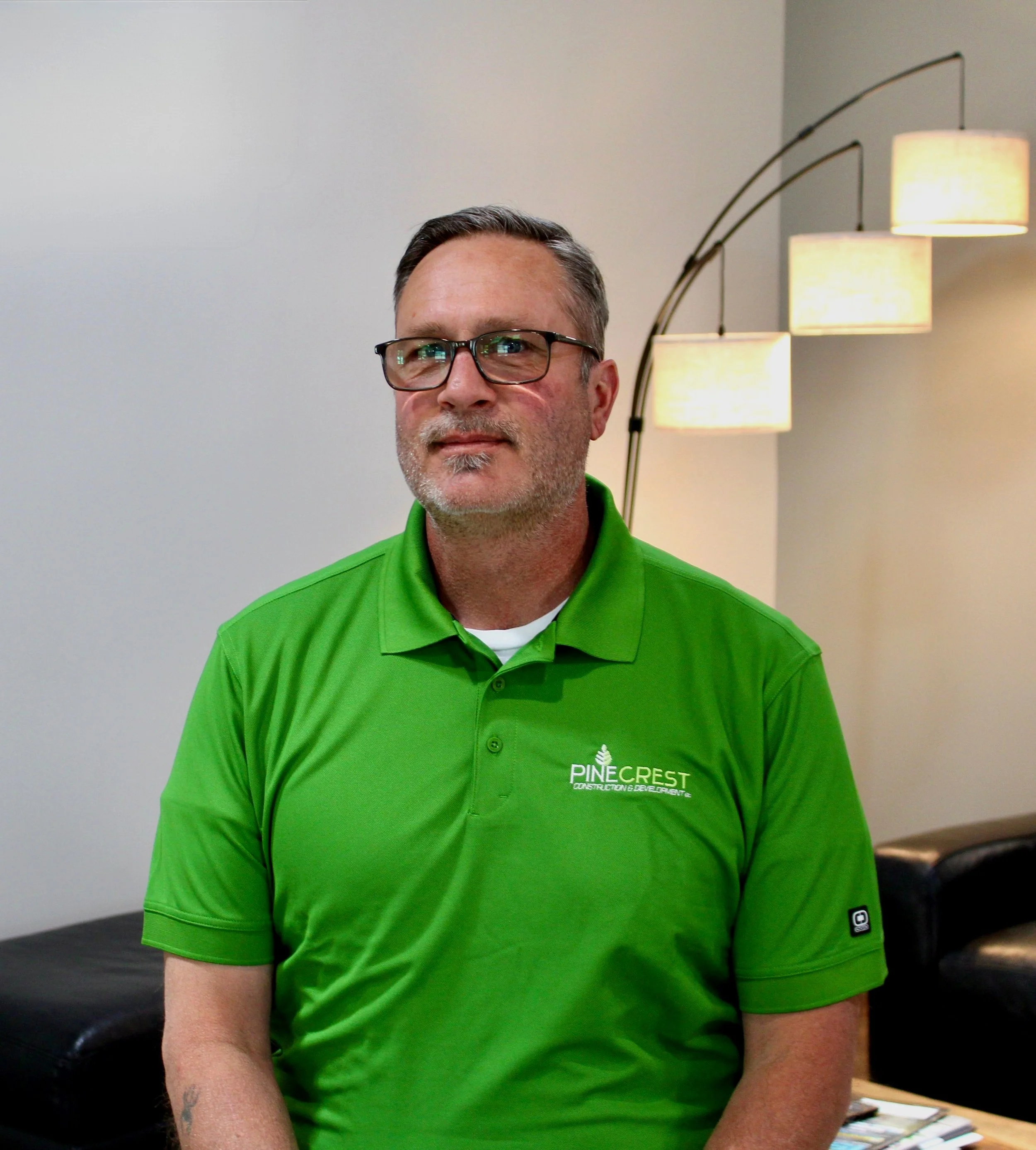 A middle-aged man with gray hair and glasses, wearing a bright green polo shirt with the logo 'Pinecrest Construction' on it, sitting in a room with modern lamps and a black leather couch.