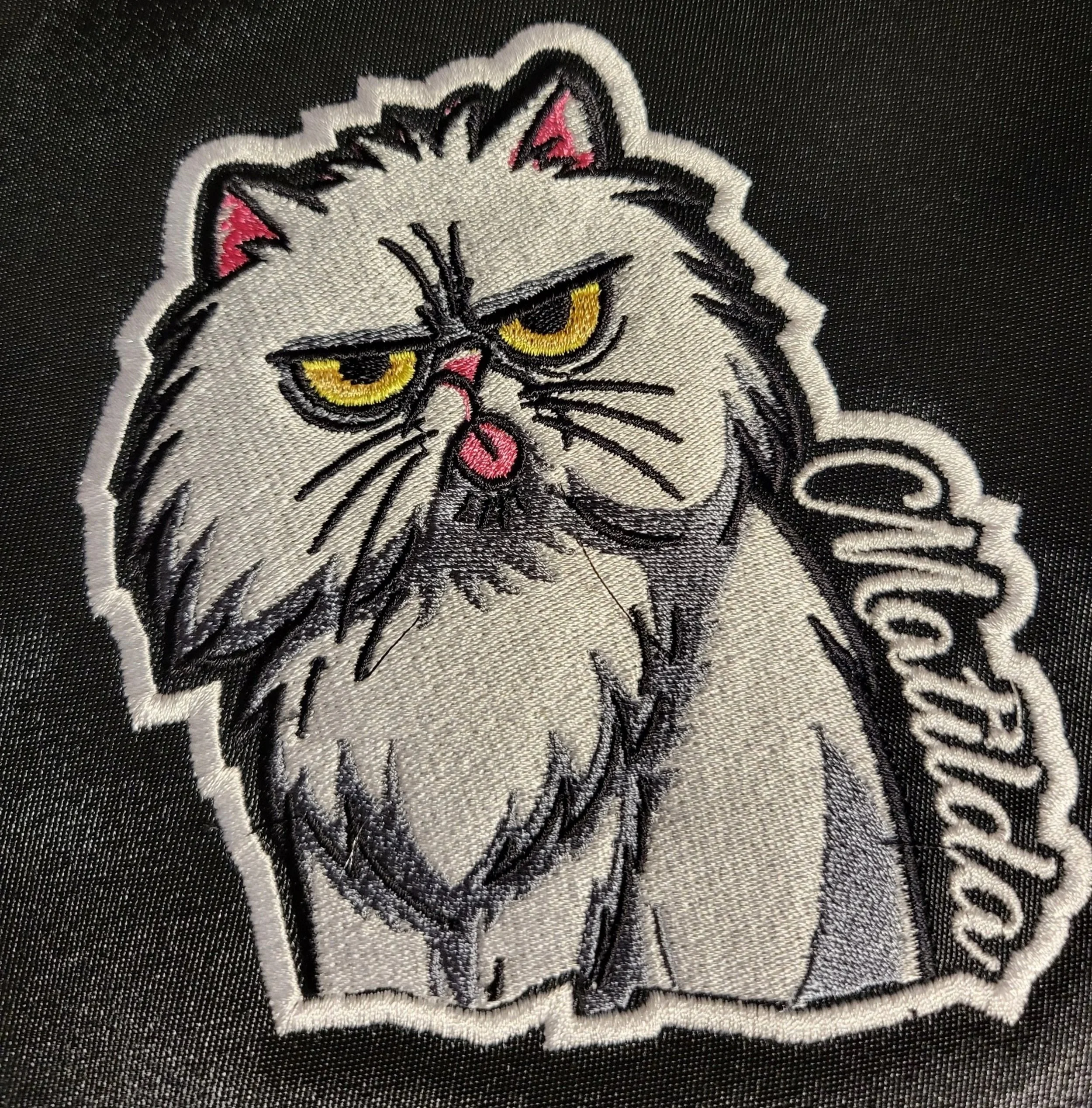 Embroidered patch of a grumpy-looking cat with yellow eyes, pink nose, and the word 'Curious' on the side.
