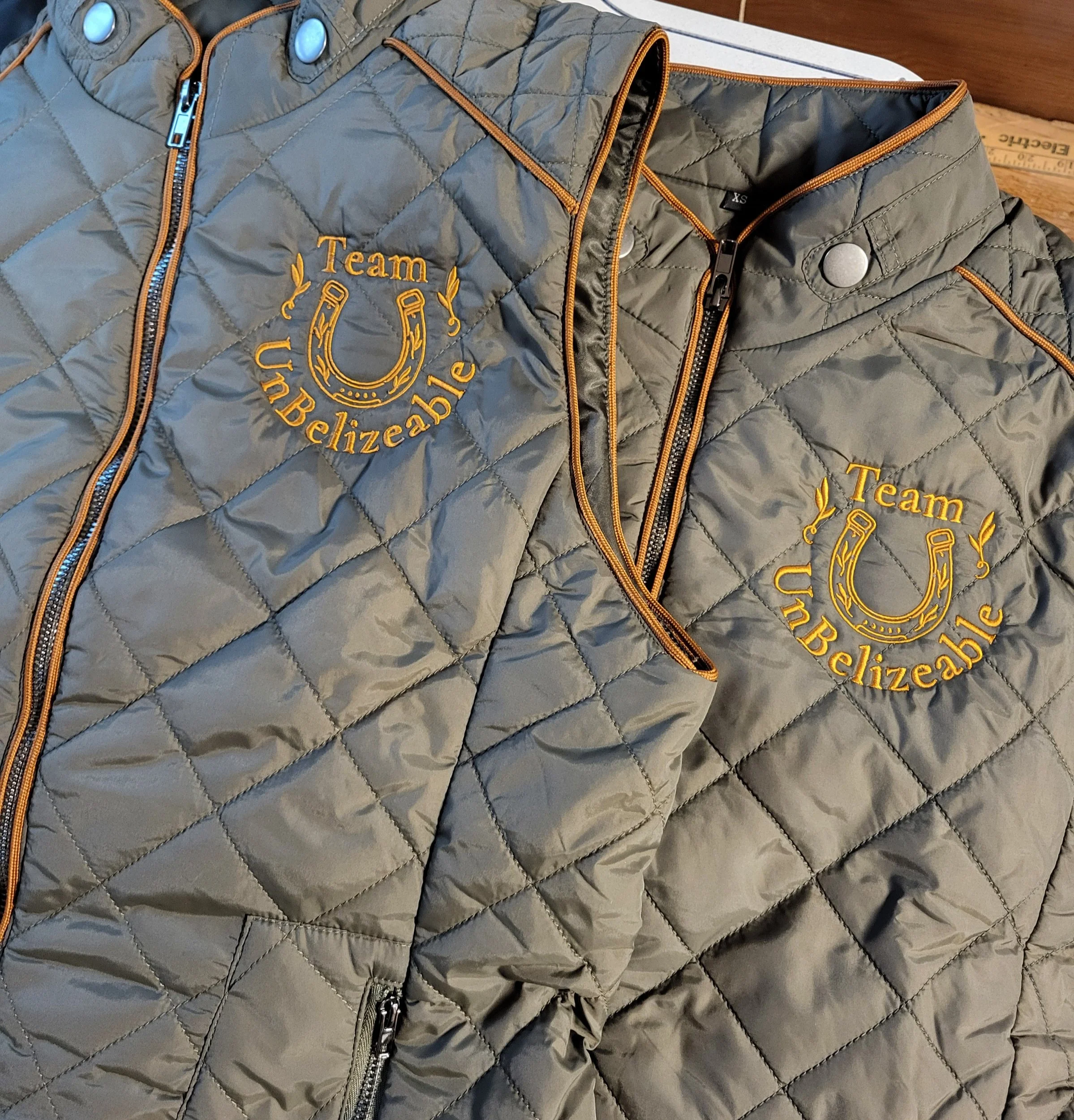Two gray quilted jackets with orange zippers and embroidery, each displaying a yellow embroidered design and text that says 'Team Unbelizable' with a horseshoe and decorative elements.