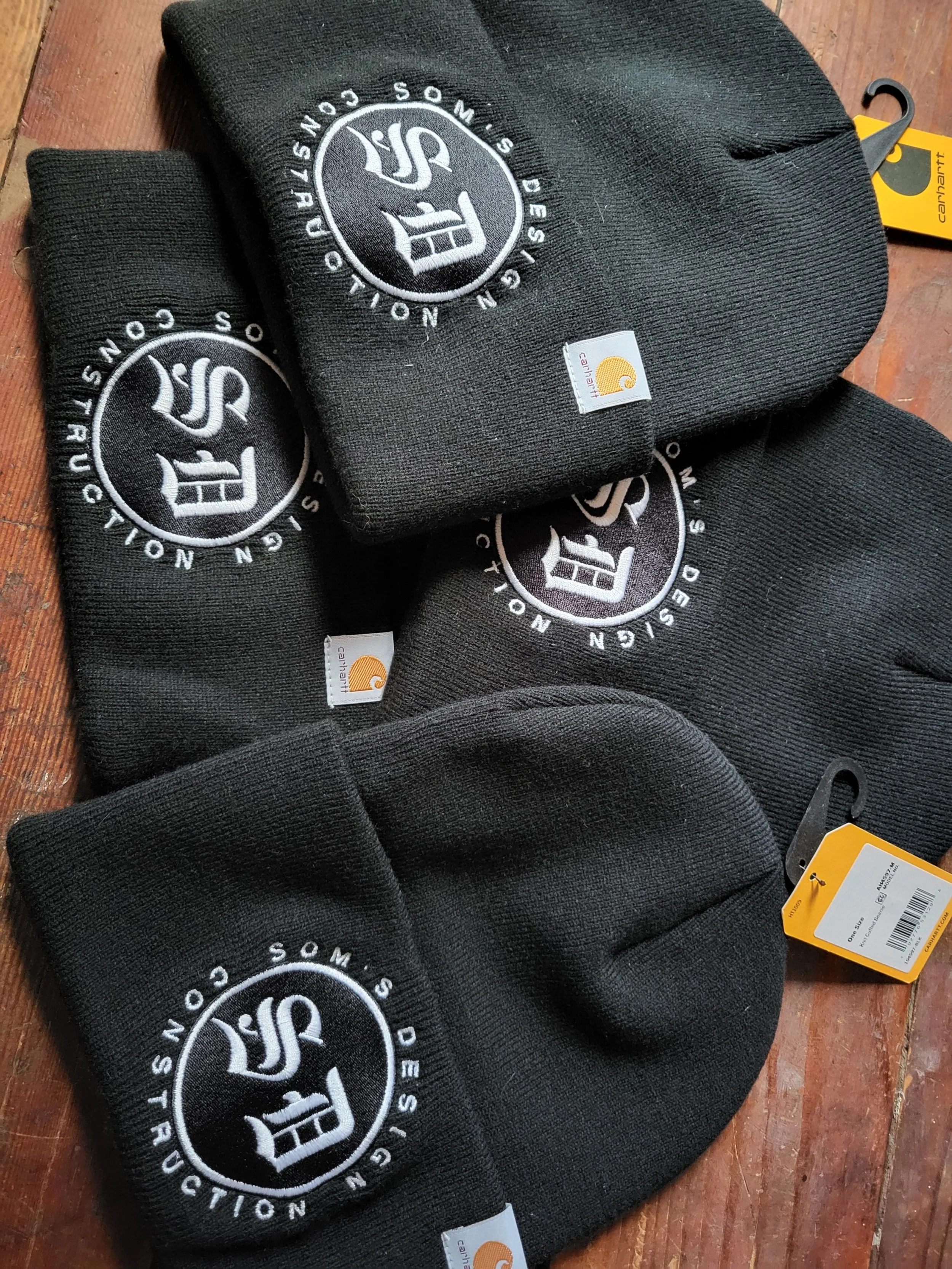 Several black Carhartt beanies with white embroidered logo and tags, placed on a wooden surface.