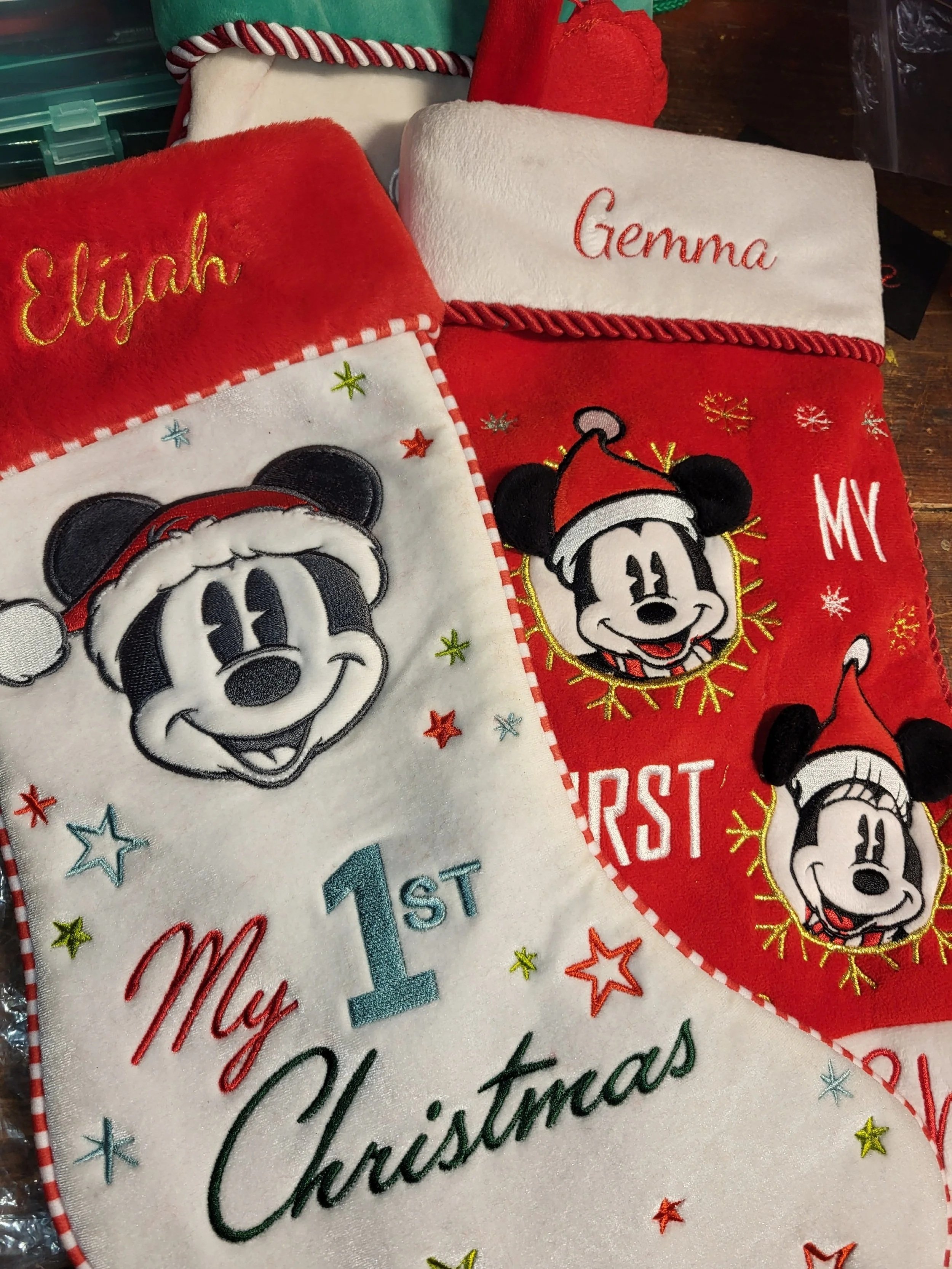 Christmas stocking decorations featuring Mickey Mouse and Minnie Mouse wearing Santa hats. One stocking is white with Mickey's face, embroidered with 'My 1st Christmas' and 'Elijah'. The other is red with Minnie and Mickey's faces, with the words 'Gemmma' and 'My First' embroidered.
