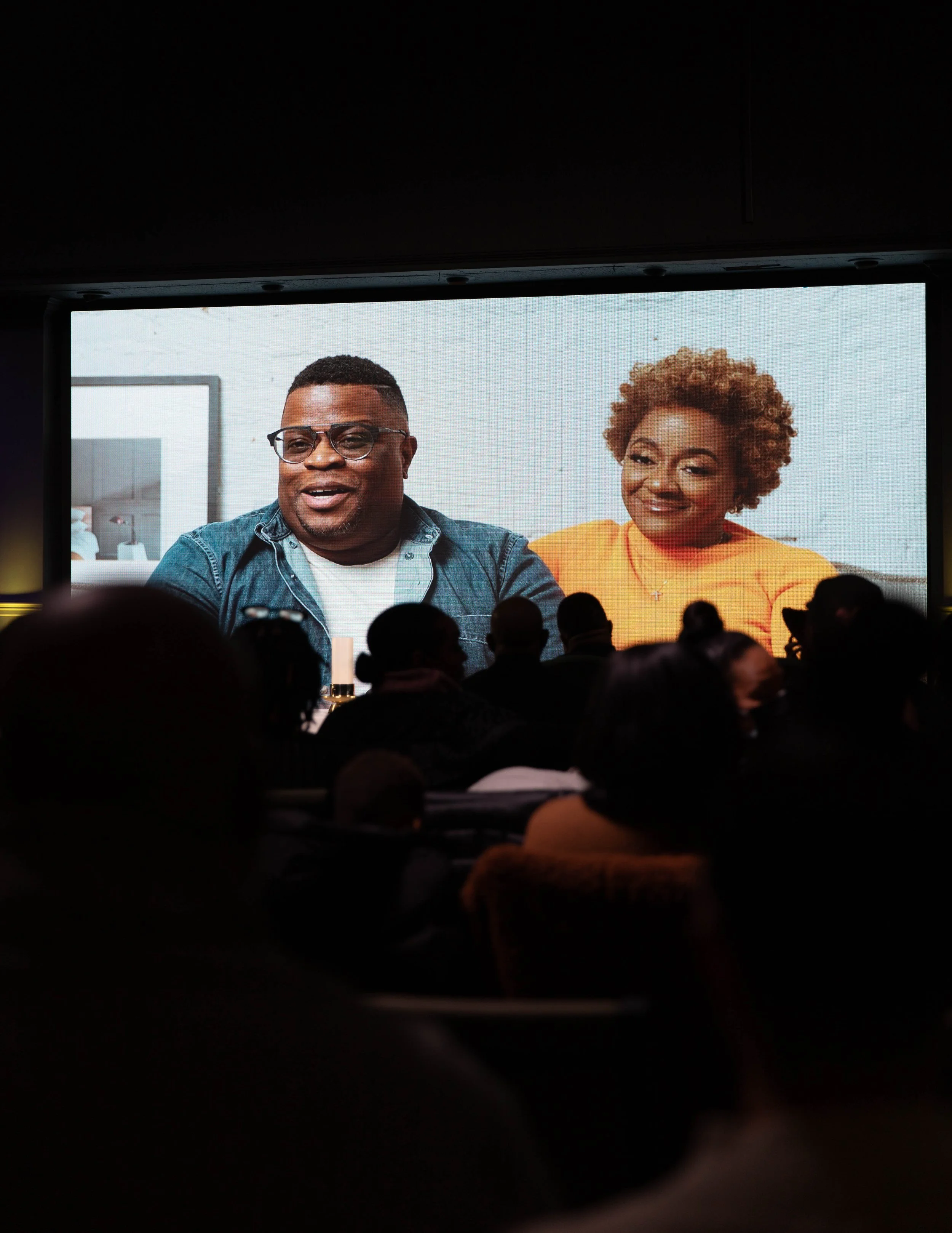 People watching a large screen showing a man and a woman speaking via video call at an indoor event.