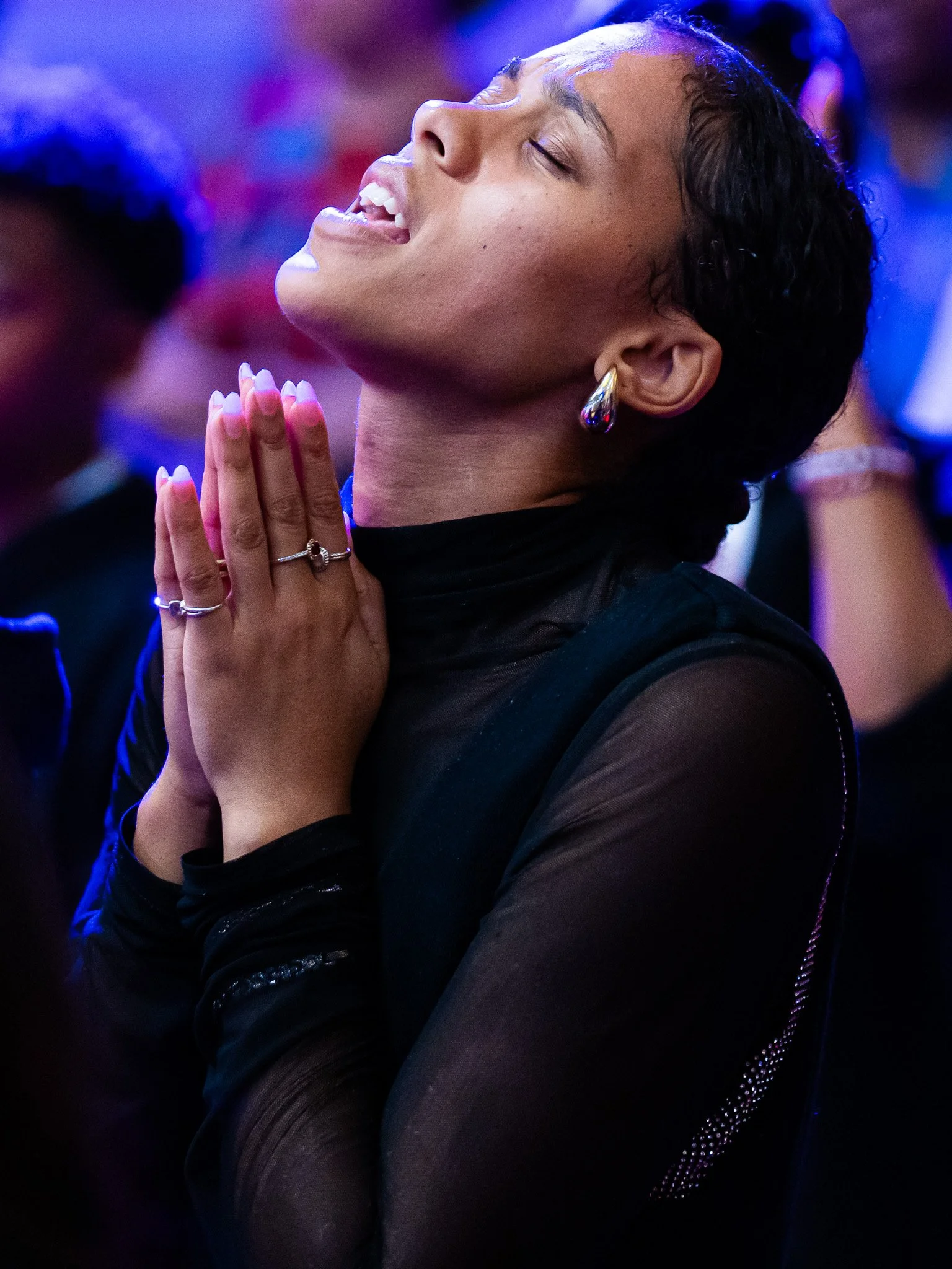 A woman with short dark hair and earrings, eyes closed, hands clasped in prayer, smiling softly in a dark, colorful setting.