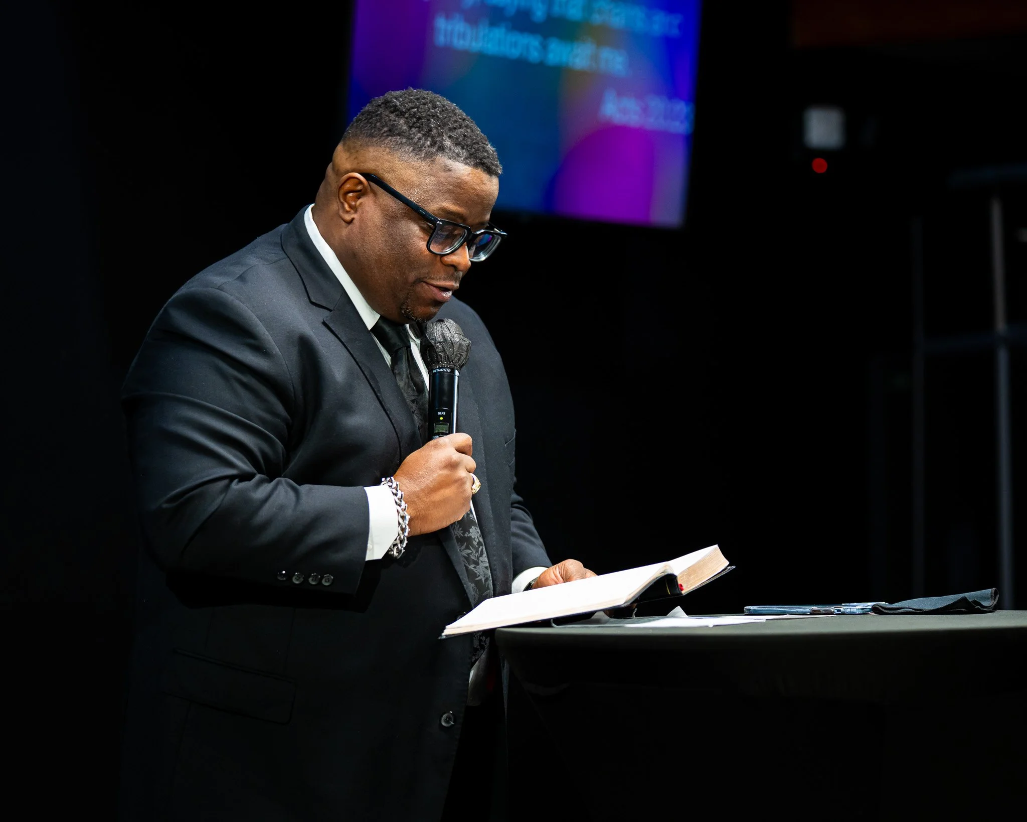 A man in a black suit and glasses is reading from a book while holding a microphone, standing at a podium with a dark background and a colorful screen in the distance.