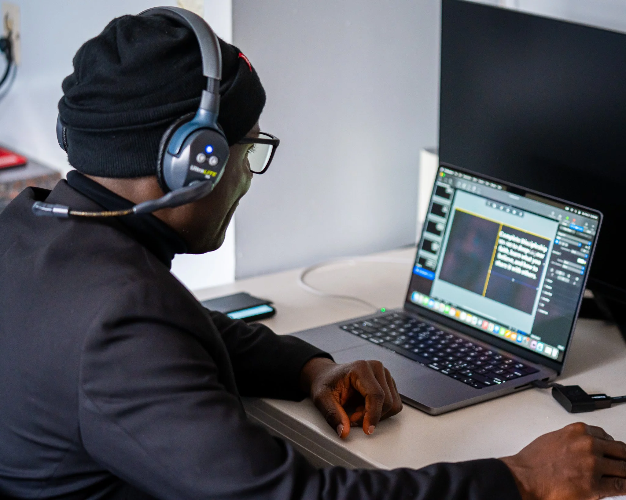 A person wearing a black beanie, glasses, and a dark jacket working at a desk with a laptop. They are wearing a headset with a microphone and appear to be editing or designing content on the laptop screen.