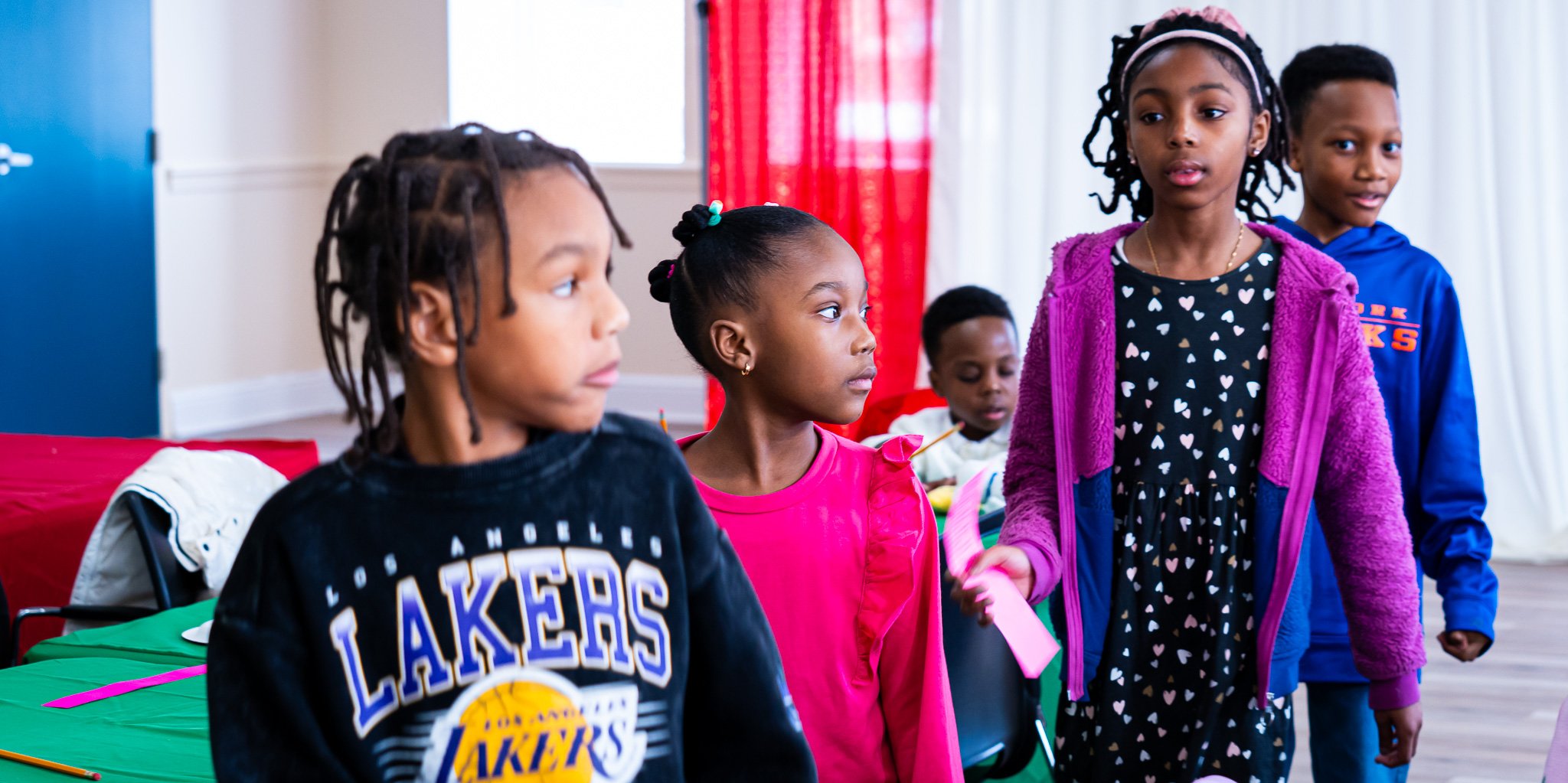Group of young children in a classroom or activity space, some holding paper and engaged in an activity.