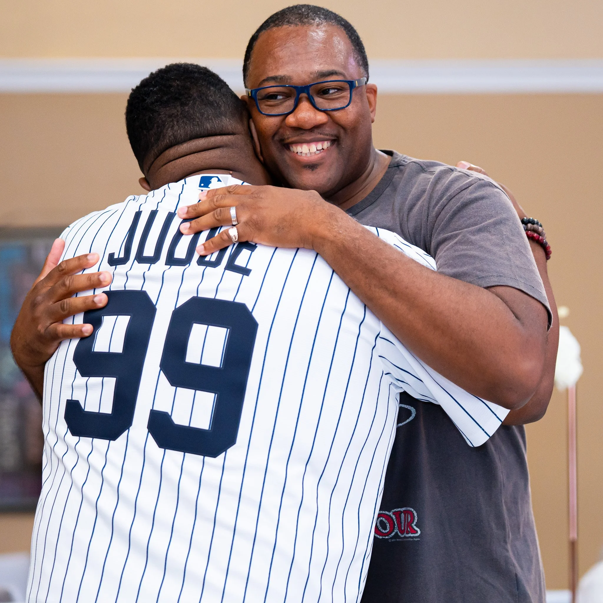 Two men embrace in a hug, one wearing a baseball jersey with the number 39 and the name 'JUDGE,' and the other smiling while wearing glasses and a gray t-shirt.