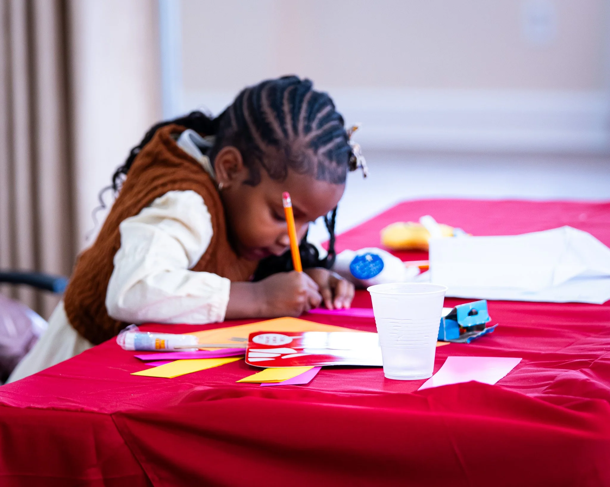A young girl with braided hair is drawing or writing on paper with a pencil at a table covered with a red tablecloth, surrounded by art supplies.
