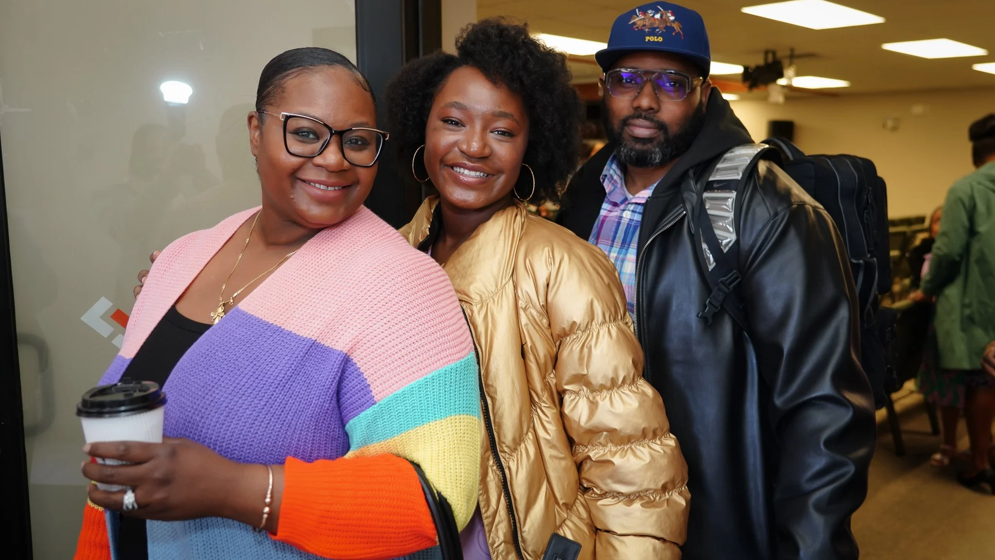Three people standing close together, smiling, in an indoor setting. The woman on the left is wearing glasses, a colorful sweater, and holding a coffee cup. The woman in the middle has curly hair and is wearing a tan jacket. The man on the right is wearing sunglasses, a cap, a leather jacket, and has a backpack.