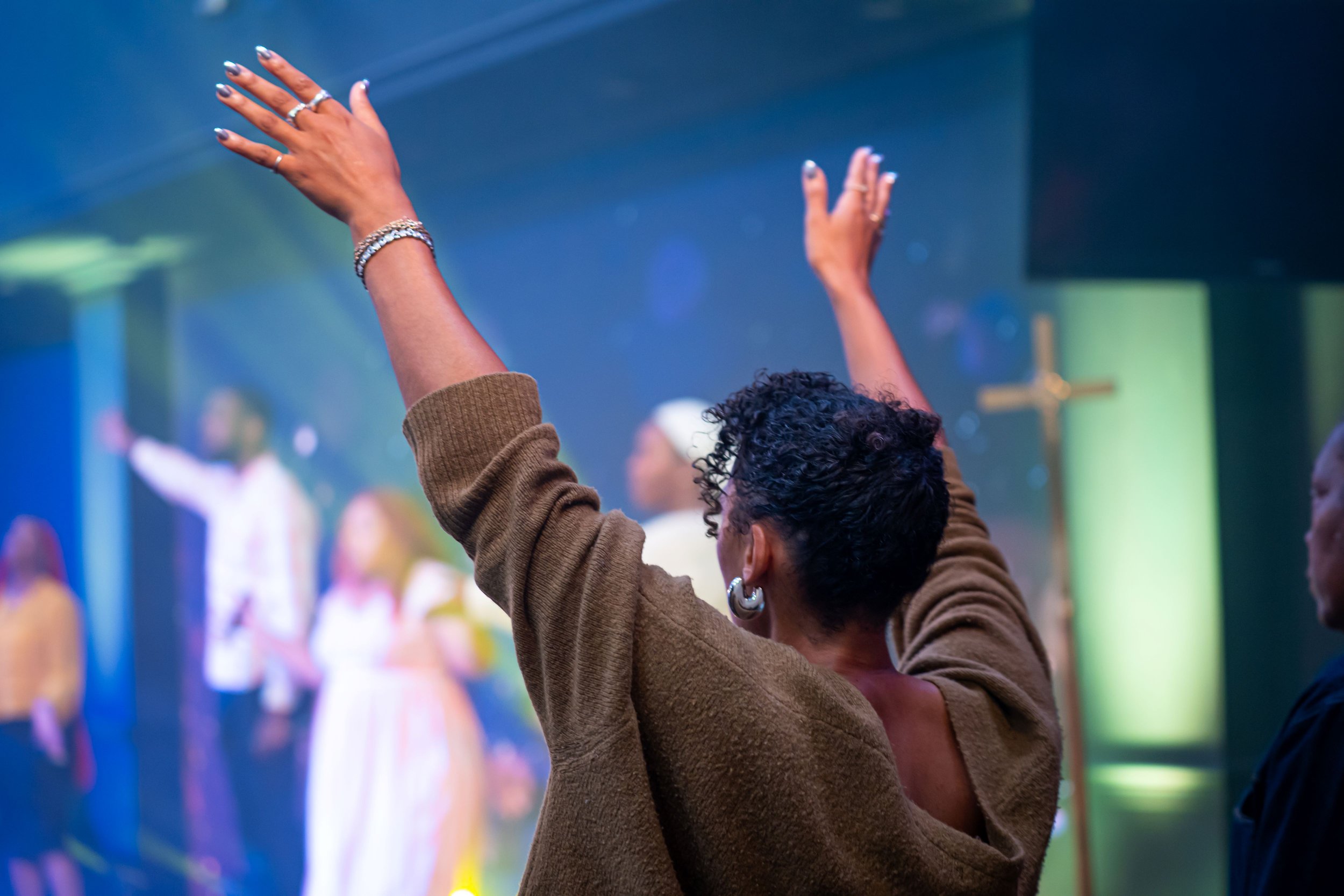 A woman with black hair and hoop earrings holding a microphone and gesturing passionately with her right arm extended outward, wearing a colorful plaid dress.