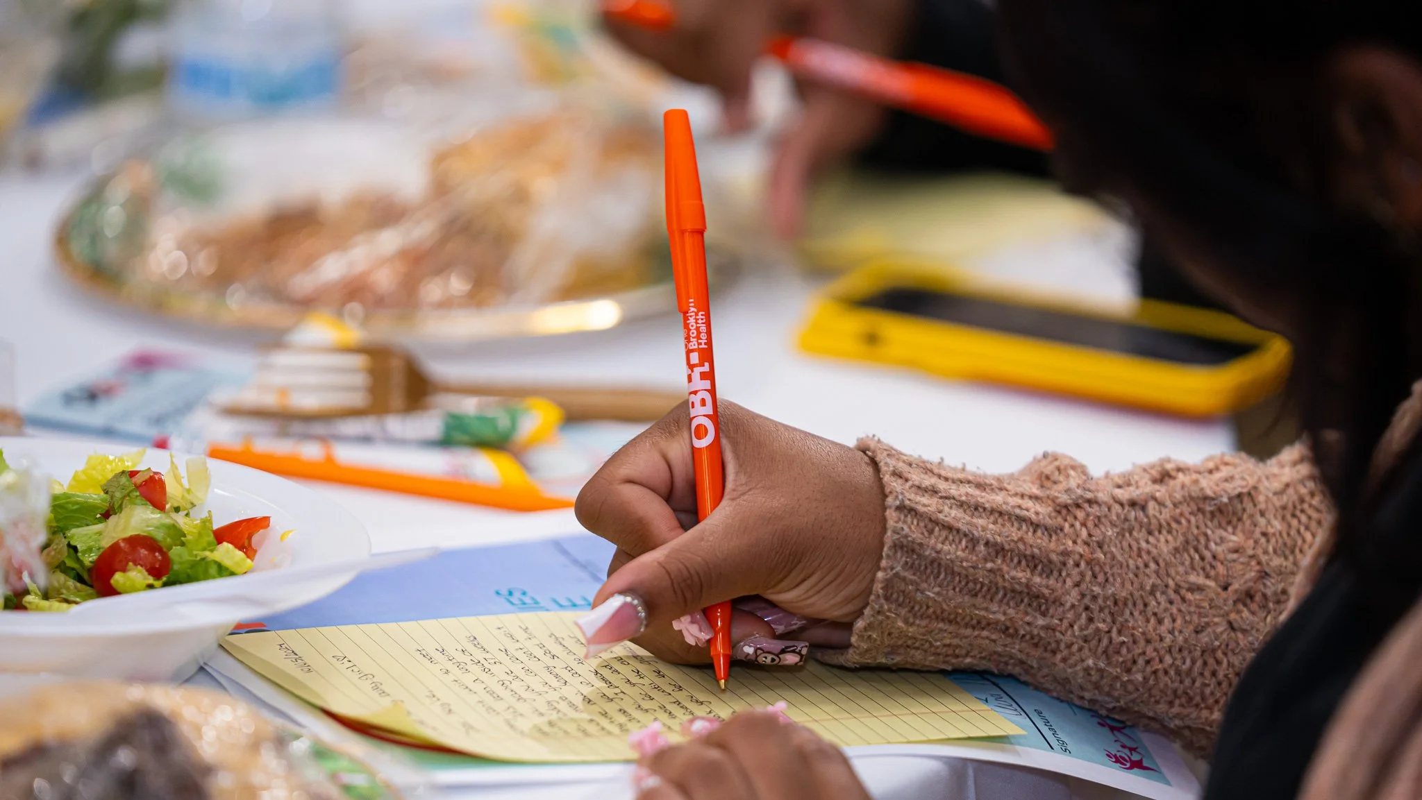 A person writing on a yellow notepad with a red pen held in their right hand, wearing a knitted pink sleeve. In the background, there is a plate of salad with lettuce and cherry tomatoes, and a blurry table with various food items and a yellow and black object.