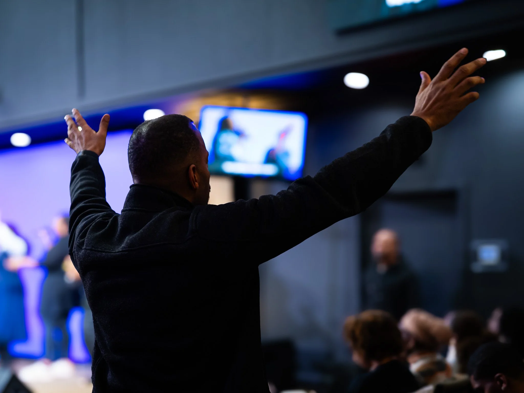 A man with short hair and dark clothing raises his hands during a presentation or event, with a large screen and several people seated in the background.