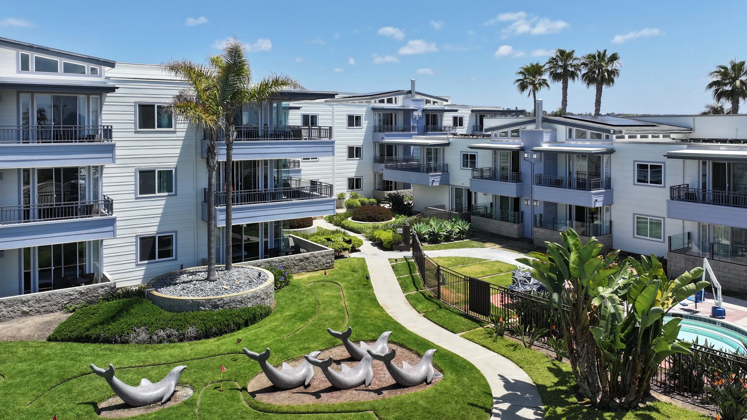 Outdoor courtyard of a modern apartment complex with white multi-story buildings, green lawns, palm trees, colorful plants, sculptures of dolphins, and a small swimming pool.