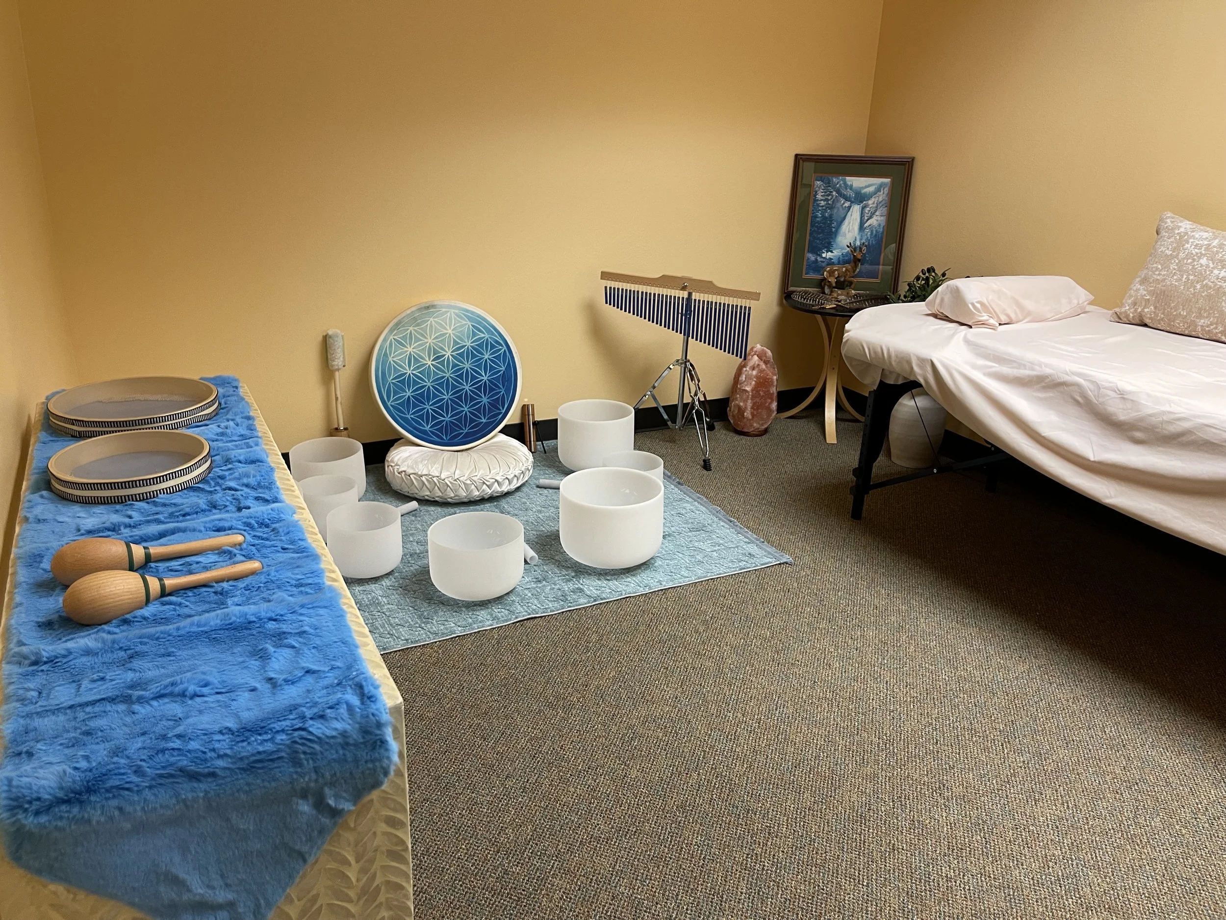 Room with musical instruments including singing bowls, ocean wave drums, a shaman drum, and a chime, along with a bed, table with decorative items, and wall art. International sound therapy office. Soundbath therapy online.