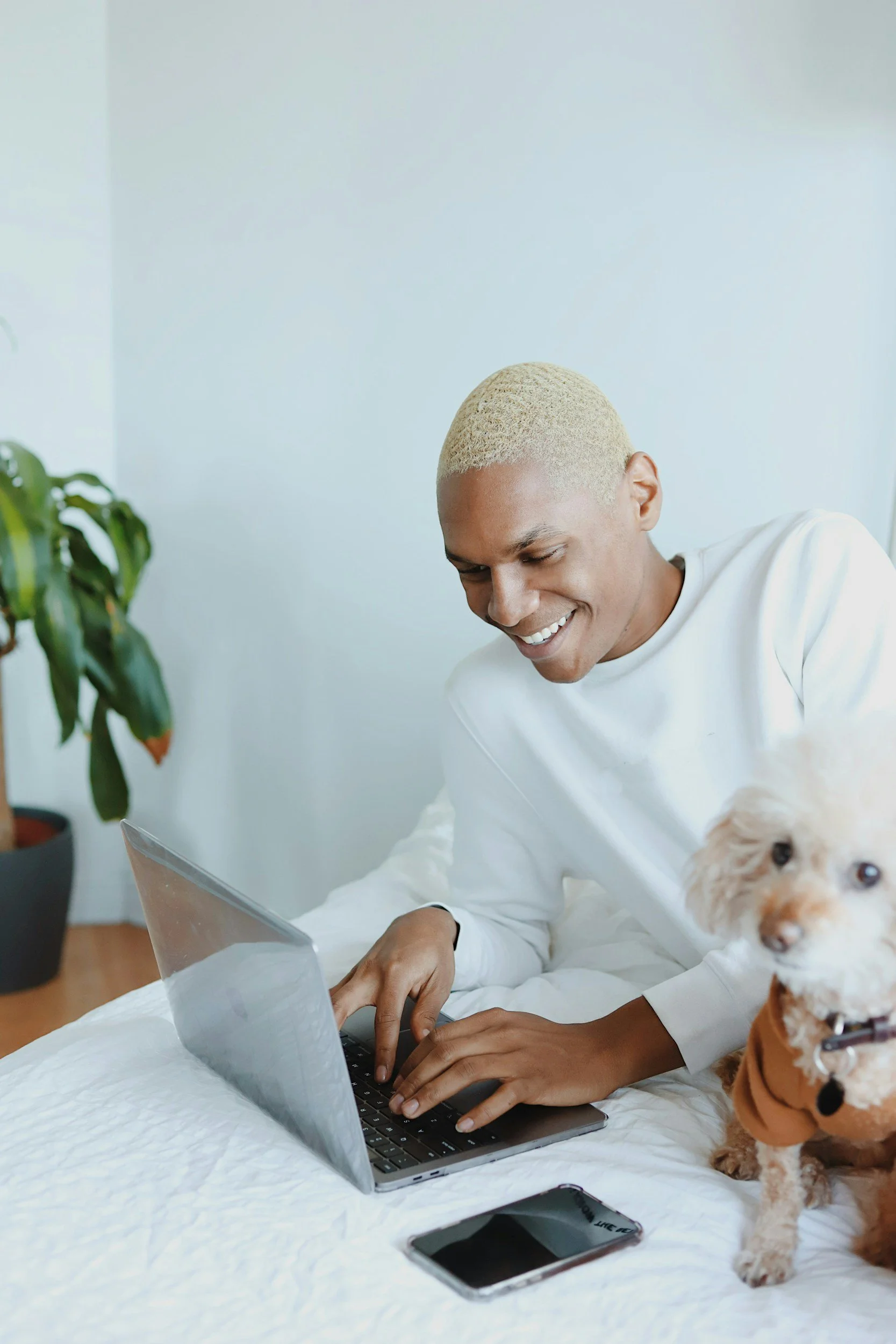 A smiling man with blonde hair working on a laptop in bed, with a small dog sitting next to him and a smartphone on the bed, in a bright room with a potted plant in the background.