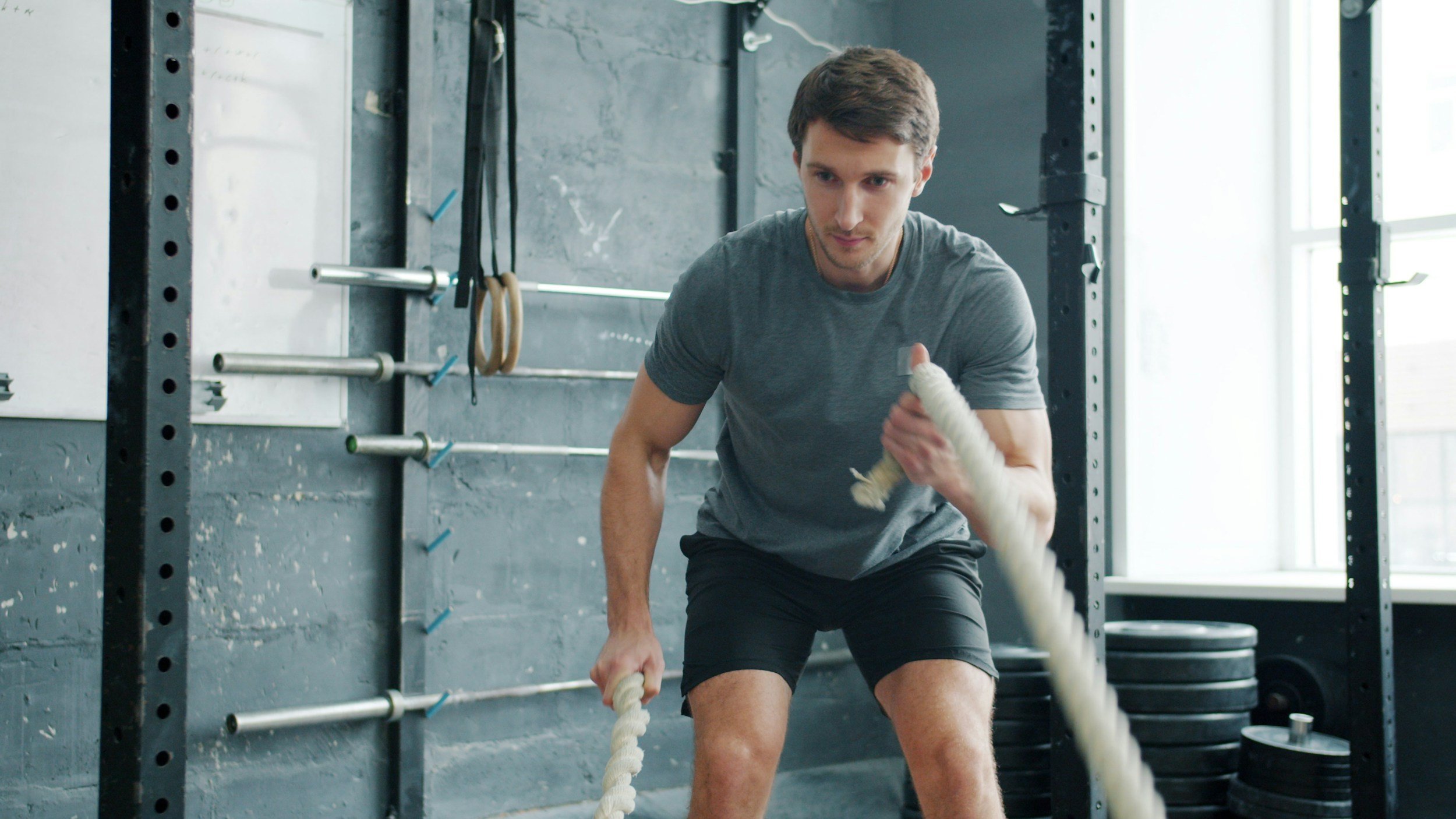 A man in a gray t-shirt and black shorts is lifting a battle rope for exercise in a gym with black weight plates and metal racks in the background.