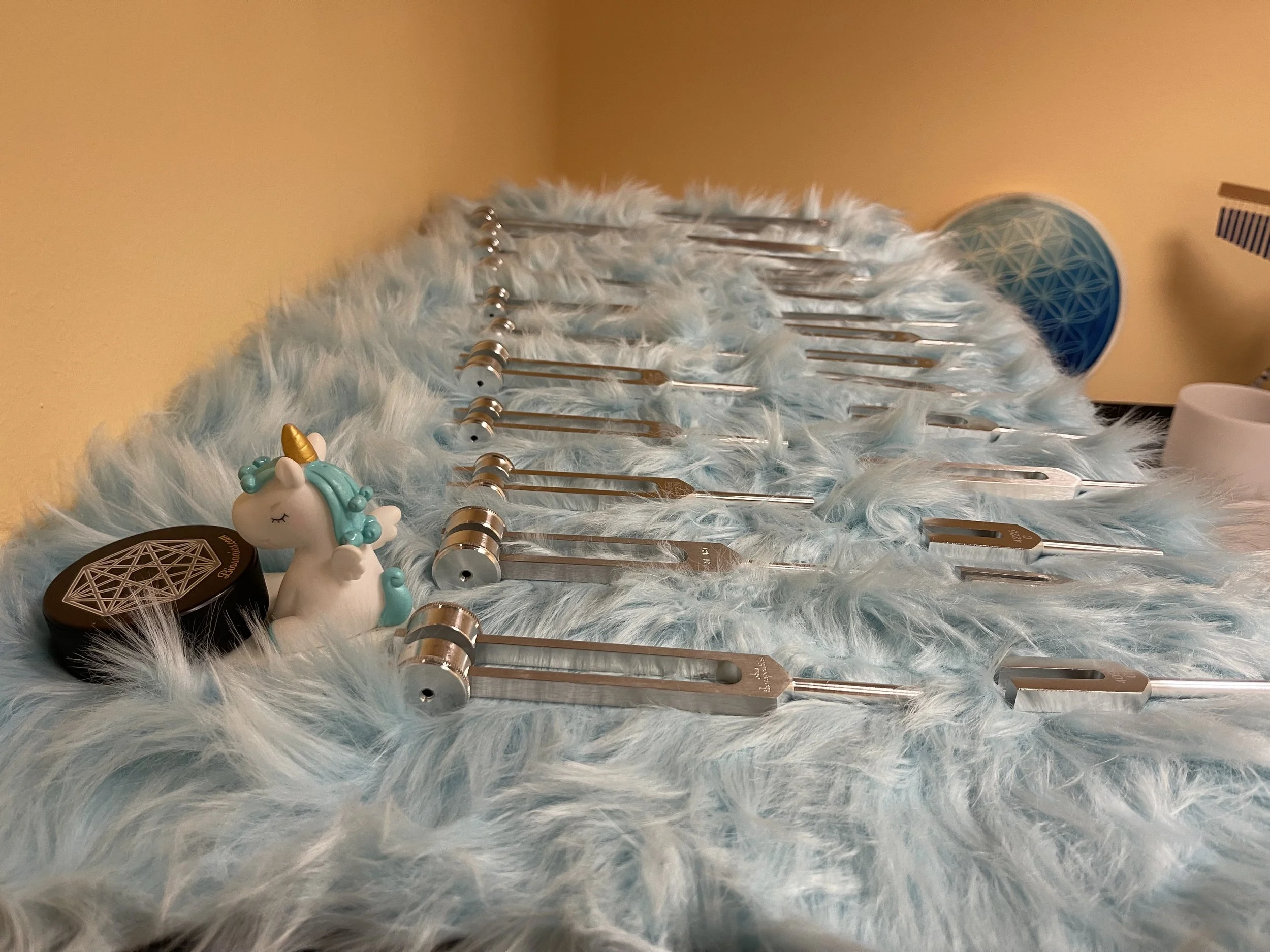 A collection of tuning forks and sound therapy tools arranged on a light blue furry surface, with a unicorn-shaped figurine and musical instruments in the background.
