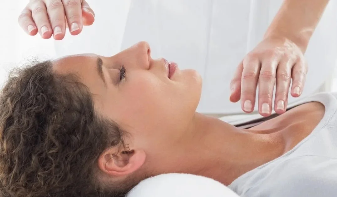 A woman receiving an energy healing treatment while lying down with her eyes closed, and a practitioner's hands hovering above her face.