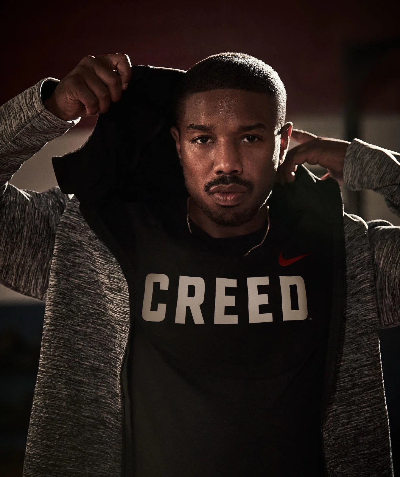 Michael B. Jordan wearing a Nike shirt with 'Creed' logo and a hoodie, looking determined.