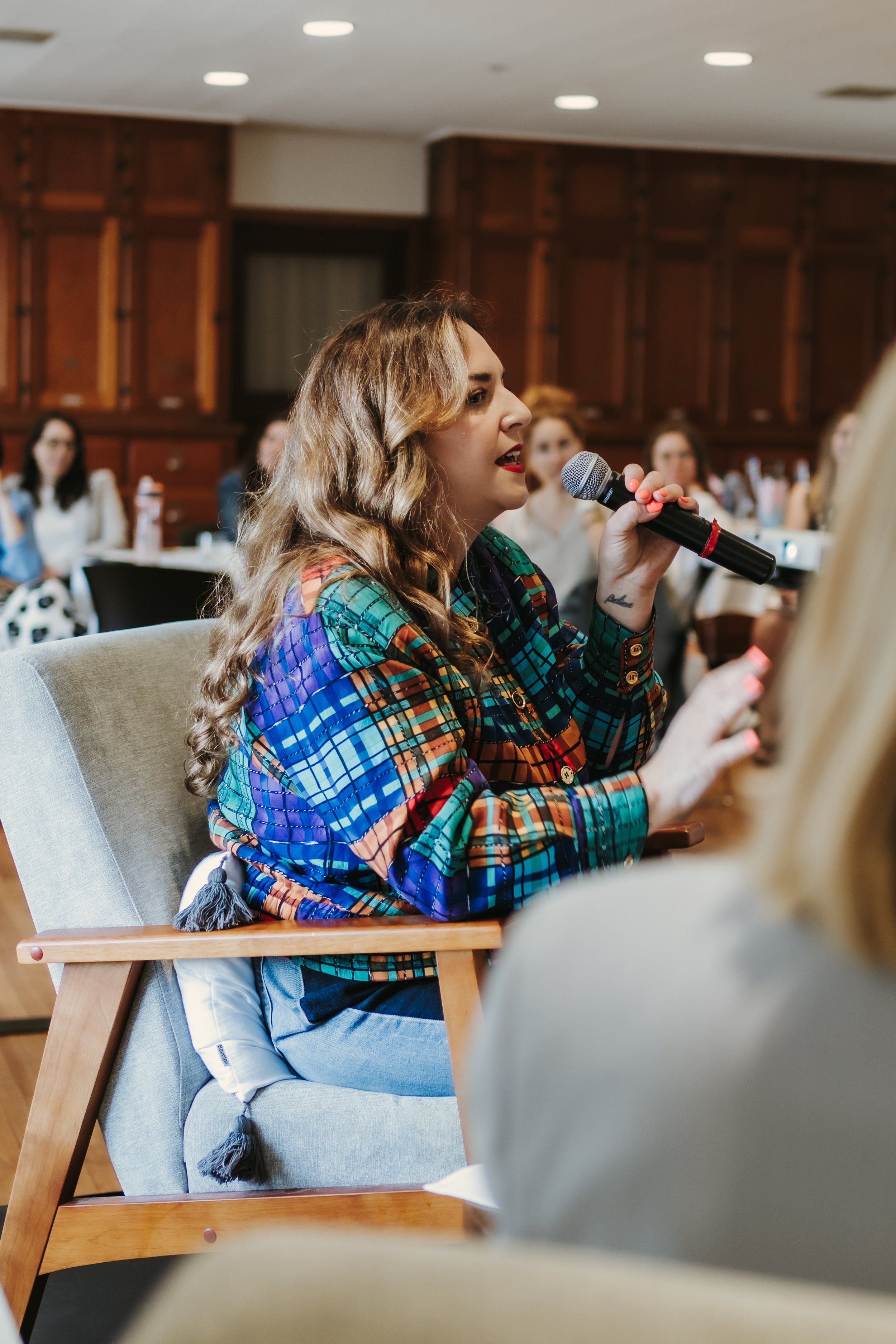 Femme parlant dans un microphone lors d'un événement, assise sur une chaise, portant une veste à motifs colorés, avec des personnes assises à l'arrière-plan.