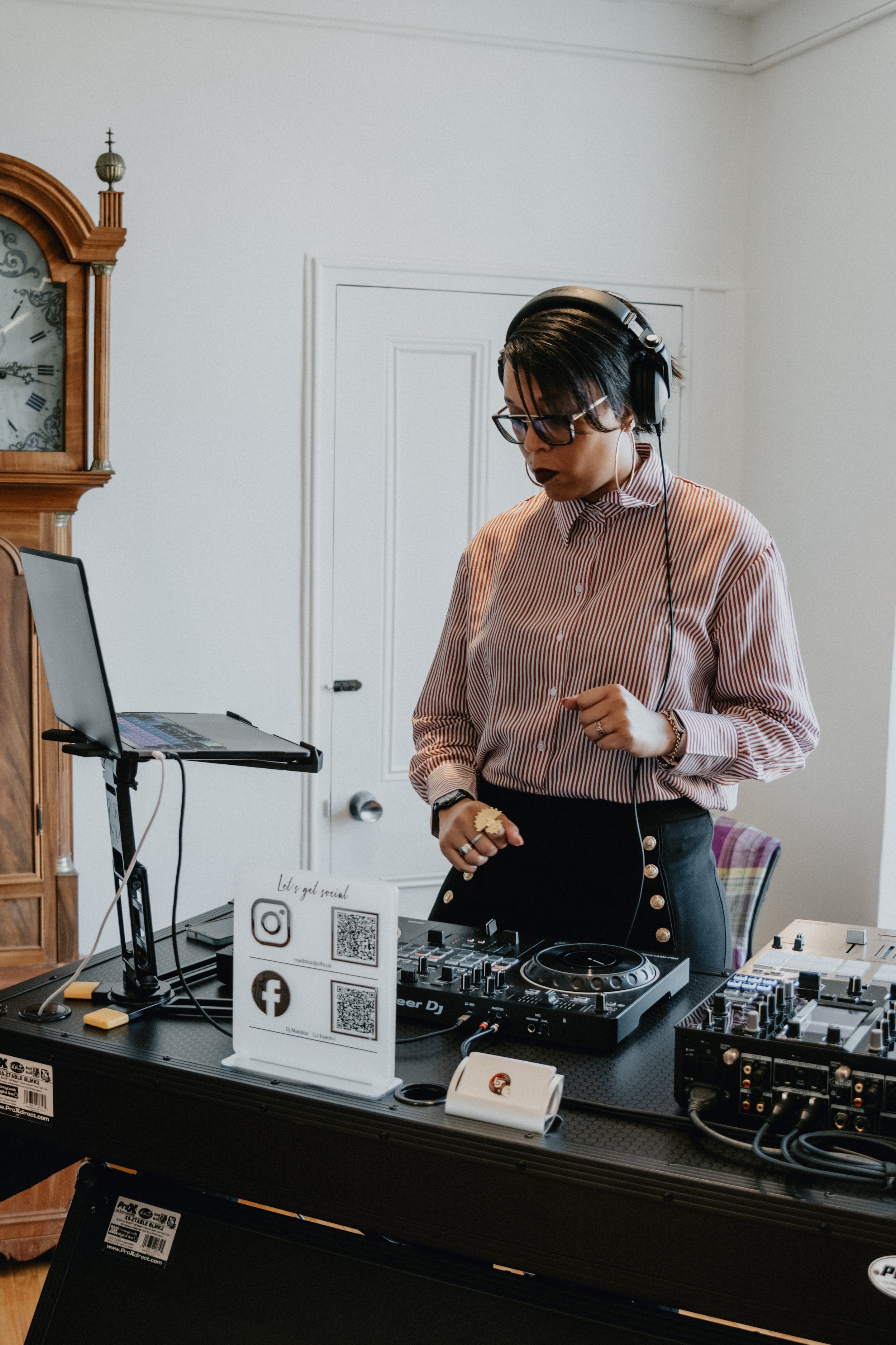DJ wearing headphones and glasses, standing behind a mixing console with a laptop. A clock and a sign with social media icons are visible.