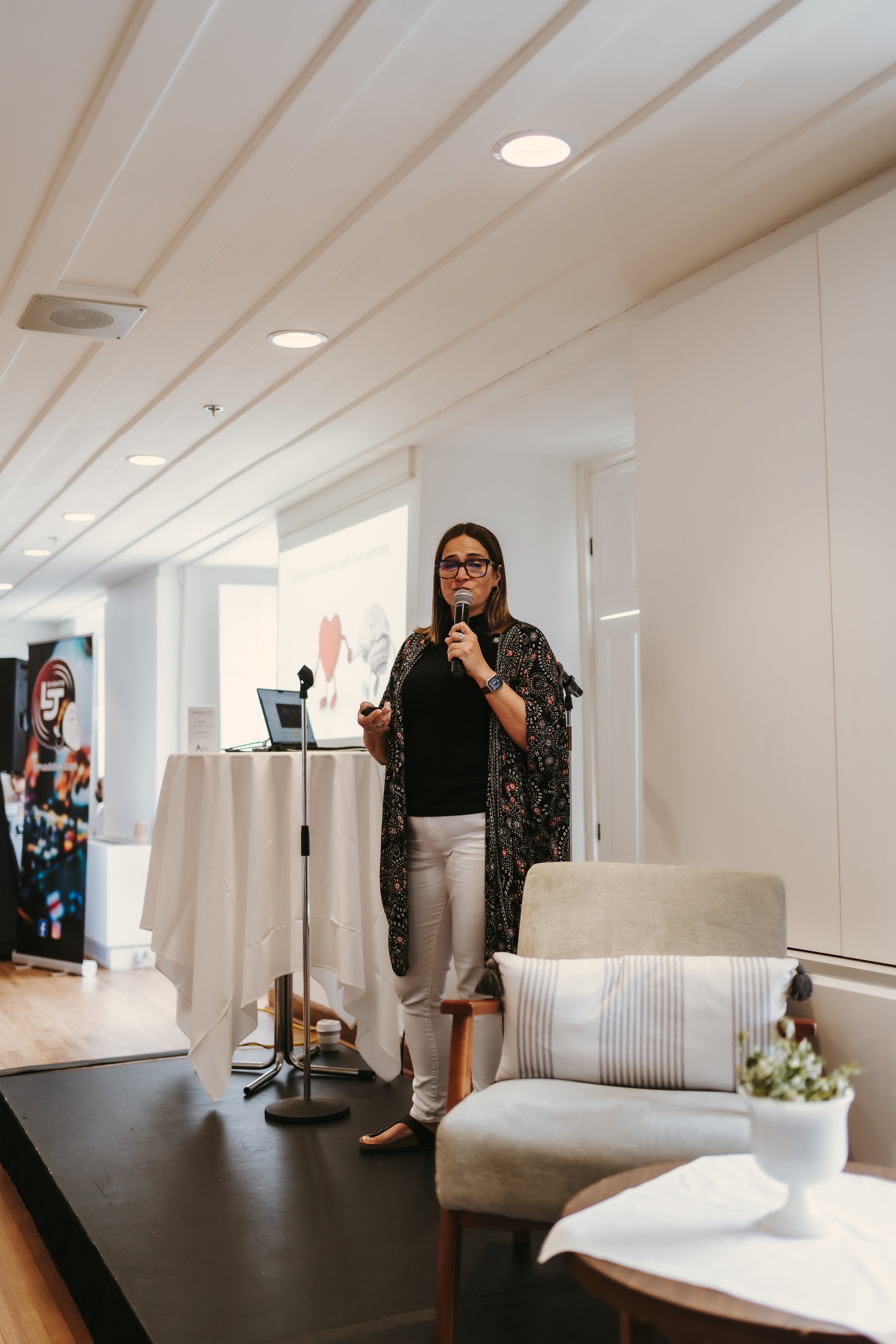 Woman giving a presentation with a microphone, standing in a conference room with projection equipment and a small table.